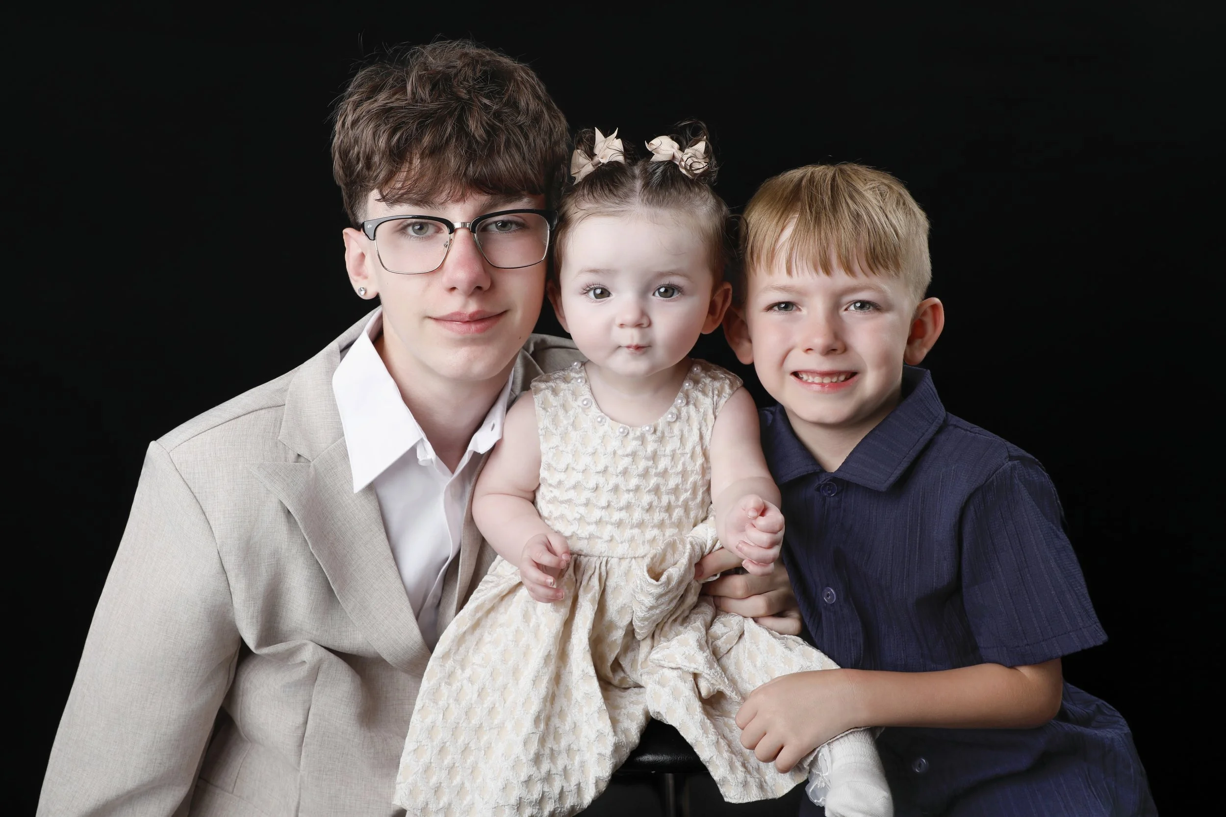 A person with short brown hair, glasses, and earrings, wearing a beige jacket over a white shirt, holds a little girl with curly hair tied in two bows, wearing a light-colored dress. A blonde boy in a dark blue shirt stands next to them, all against 