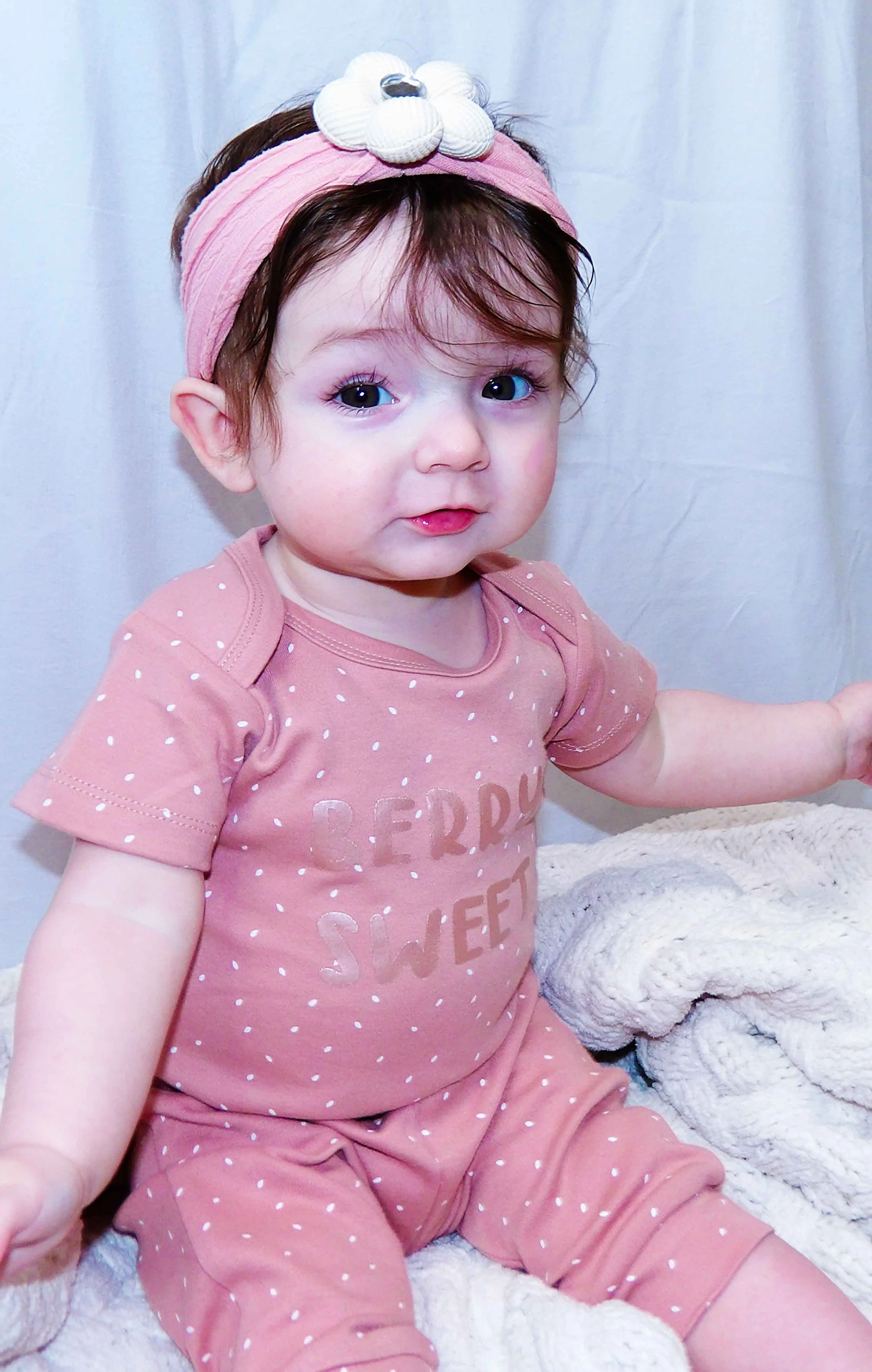 A young girl with dark hair wearing a pink headband with a white flower decoration, pink polka dot pajamas, sitting on a soft blanket, looking at the camera with a gentle expression.