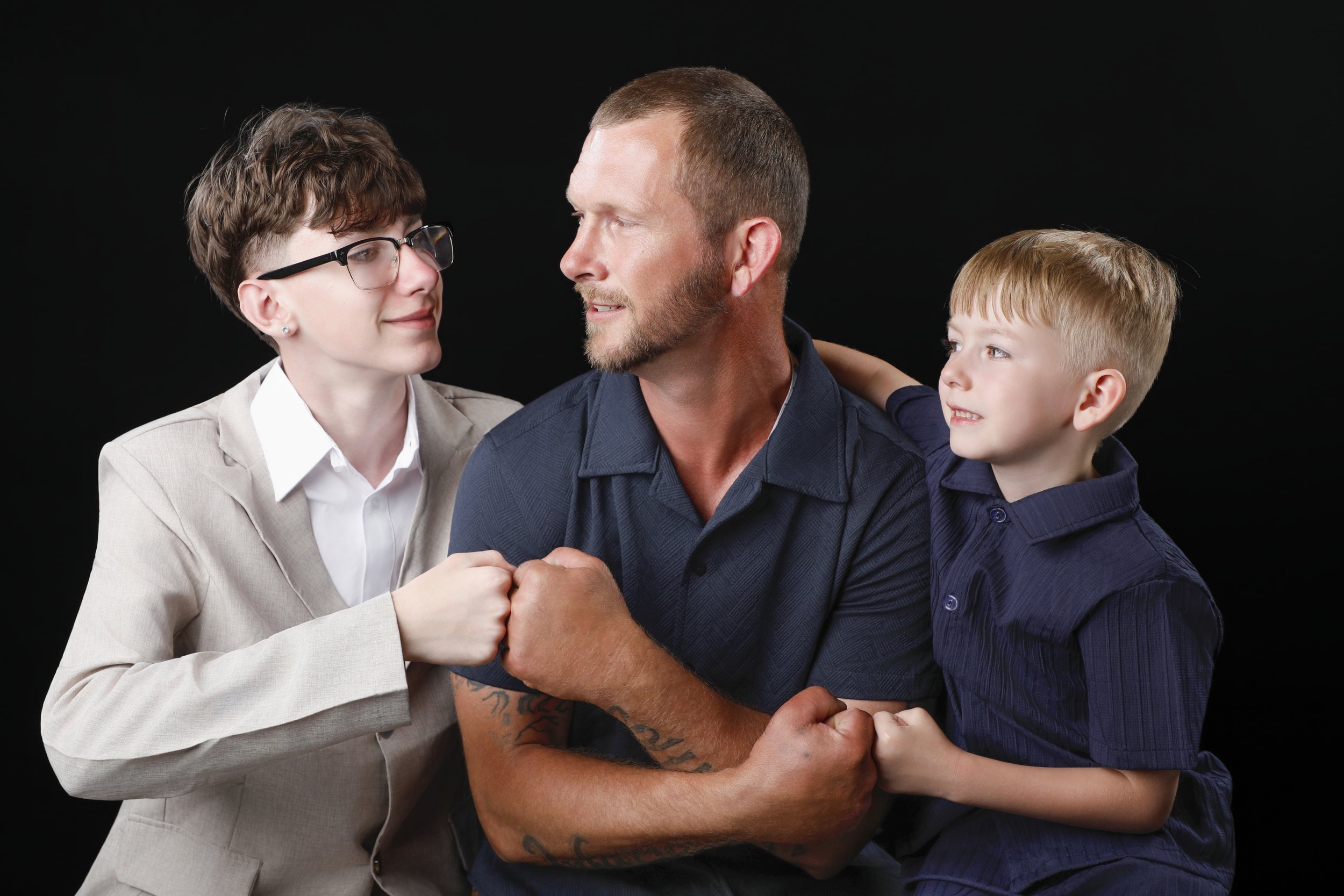 A man with a beard and a blue polo shirt, a woman with short brown hair and glasses, and a young boy with short blond hair, all clasping hands together in a show of unity against a black background.
