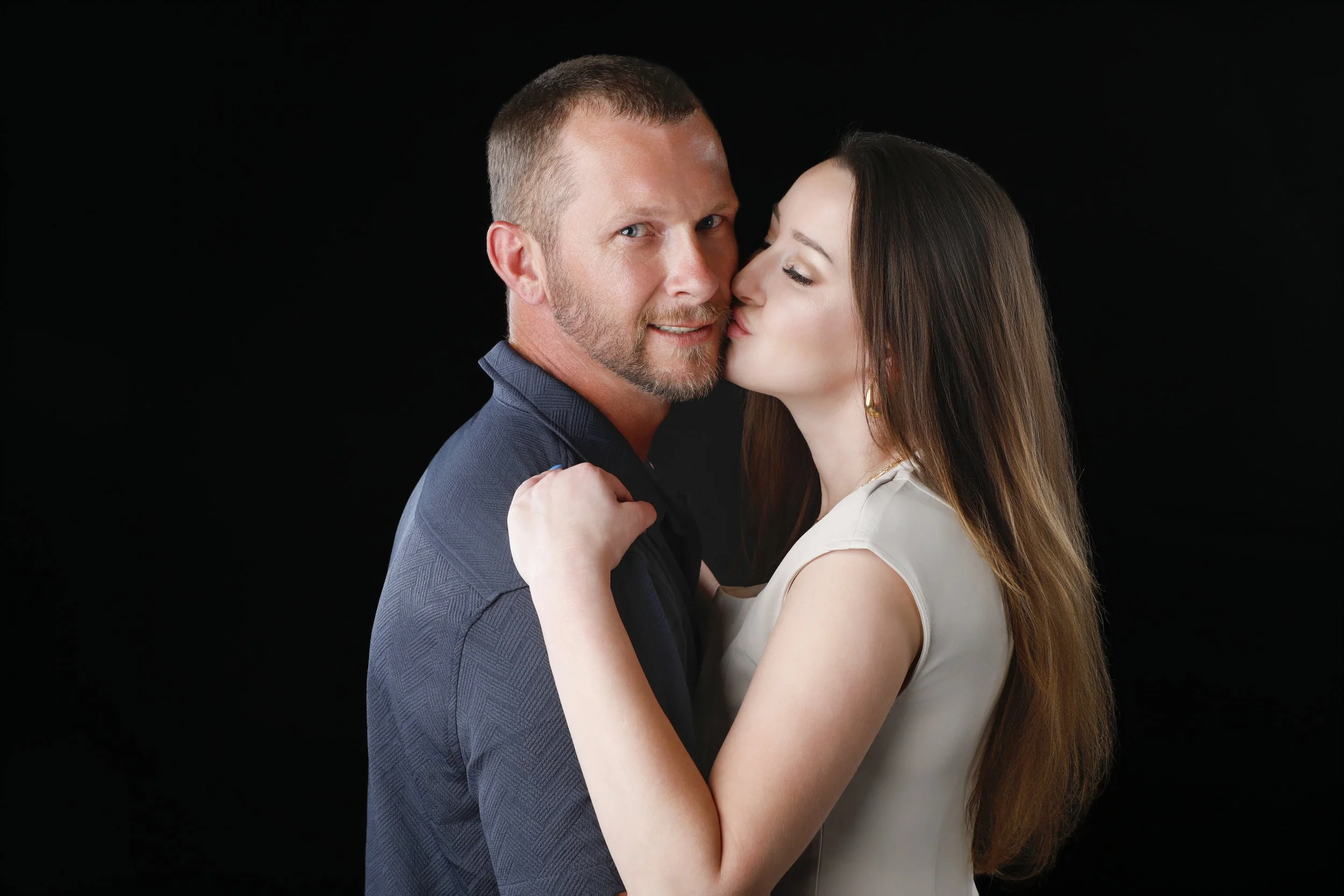 A man with short hair and a beard being kissed on the cheek by a woman with long brown hair, both against a black background.