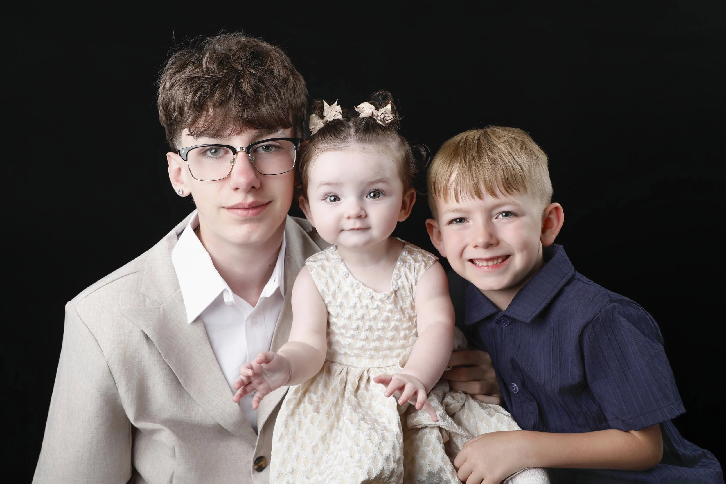 A young adult male with glasses and brown hair, a young girl with curly hair styled with bows, and a young boy with blond hair, all smiling and sitting close together against a black background.