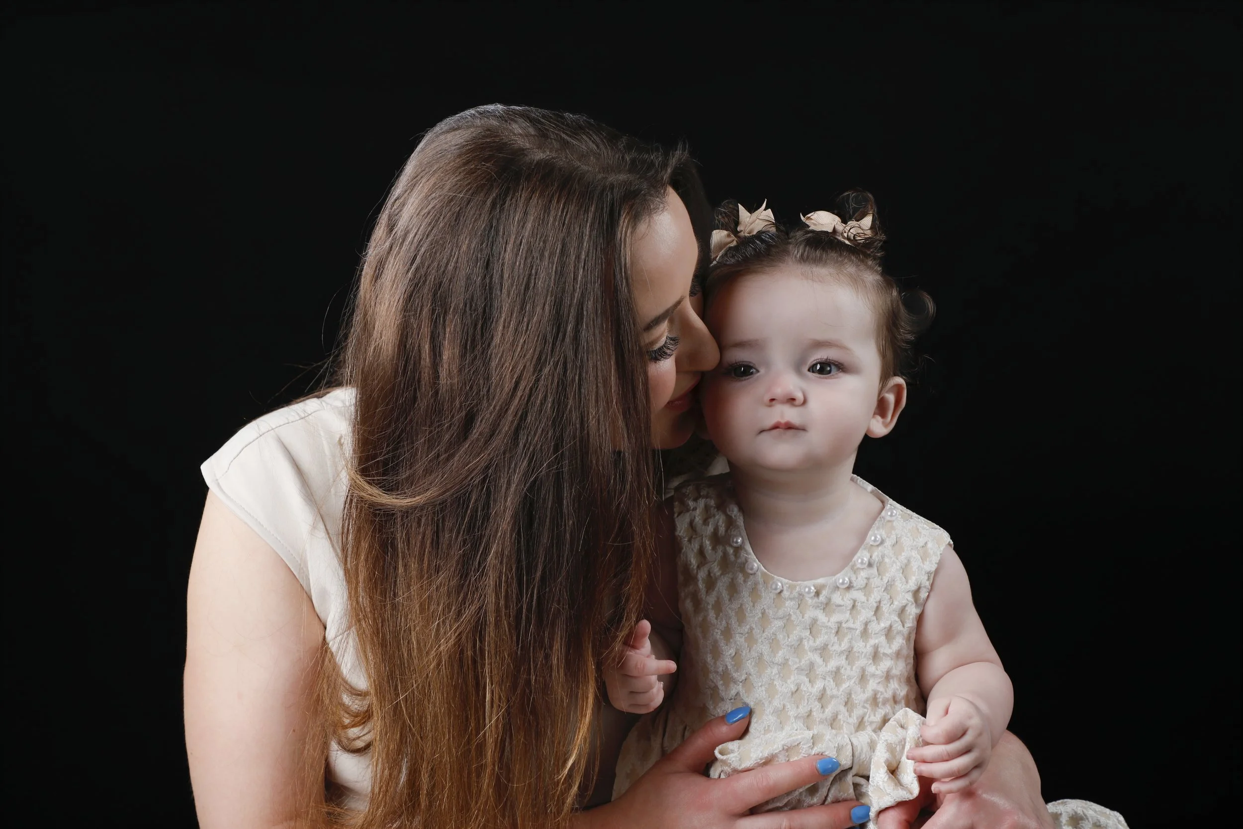 A woman with long brown hair holding a young girl with curly hair and beige bows, against a black background.