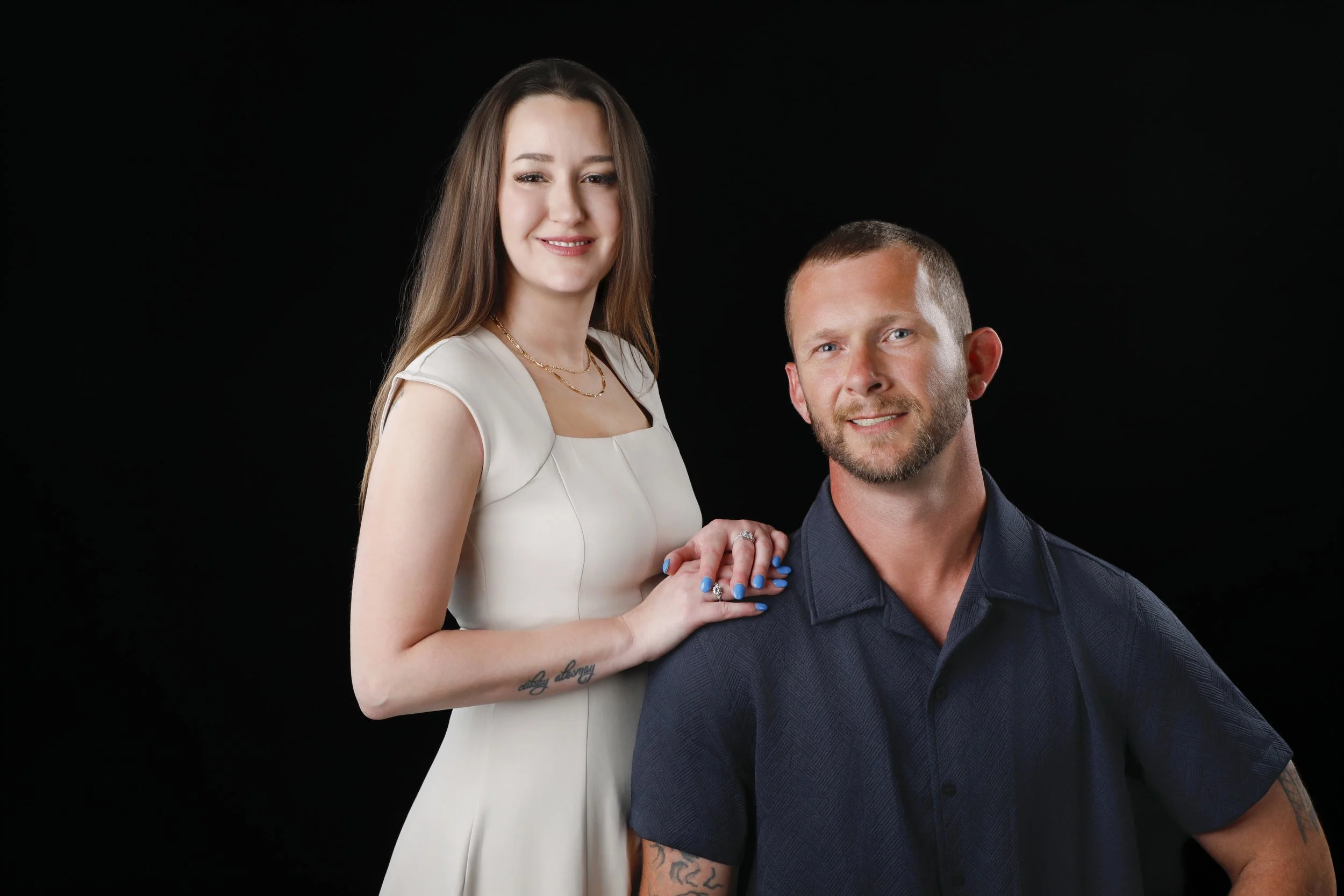 A woman with long brown hair, dressed in a white dress, holding the hand of a man with short brown hair and a beard, wearing a dark shirt, both posed against a black background.