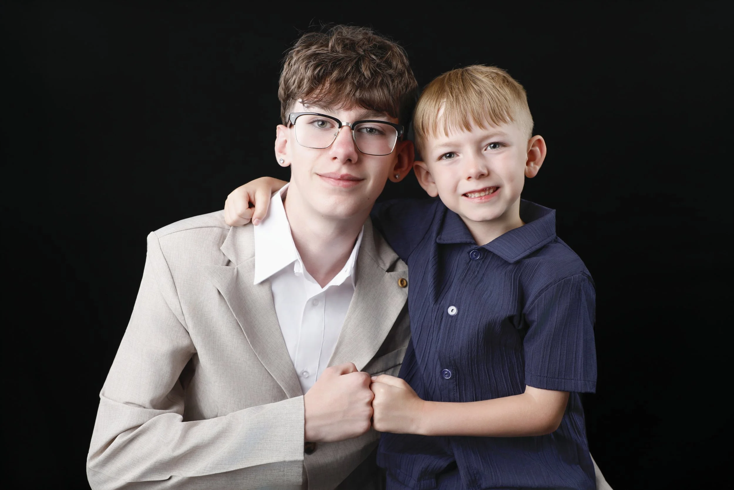 A woman and a young boy are smiling and holding hands, with the boy's arm around the woman's shoulder, against a black background.