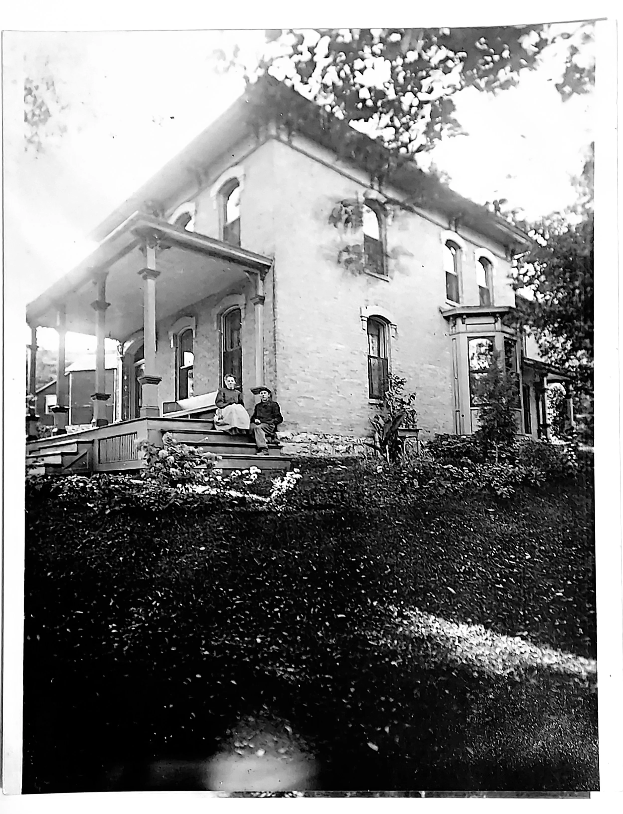 Black and white historical photograph of a two-story house with a porch, a man and woman seated on the porch steps in front of the house