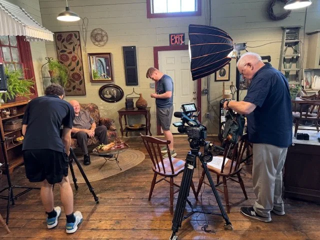 Film crew setting up equipment inside a historic downtown building with an local resident sitting on a sofa, while another crew member adjusts a camera, and a person stands near a light with a large umbrella.