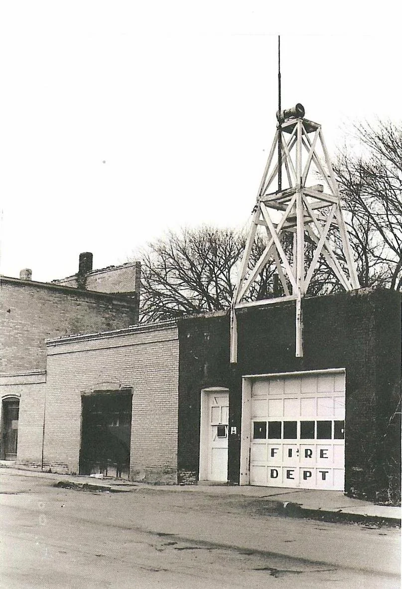 An old fire department building with a sign that says 'FIRE DEPT' on the garage door, and a tall fire watchtower on the roof.