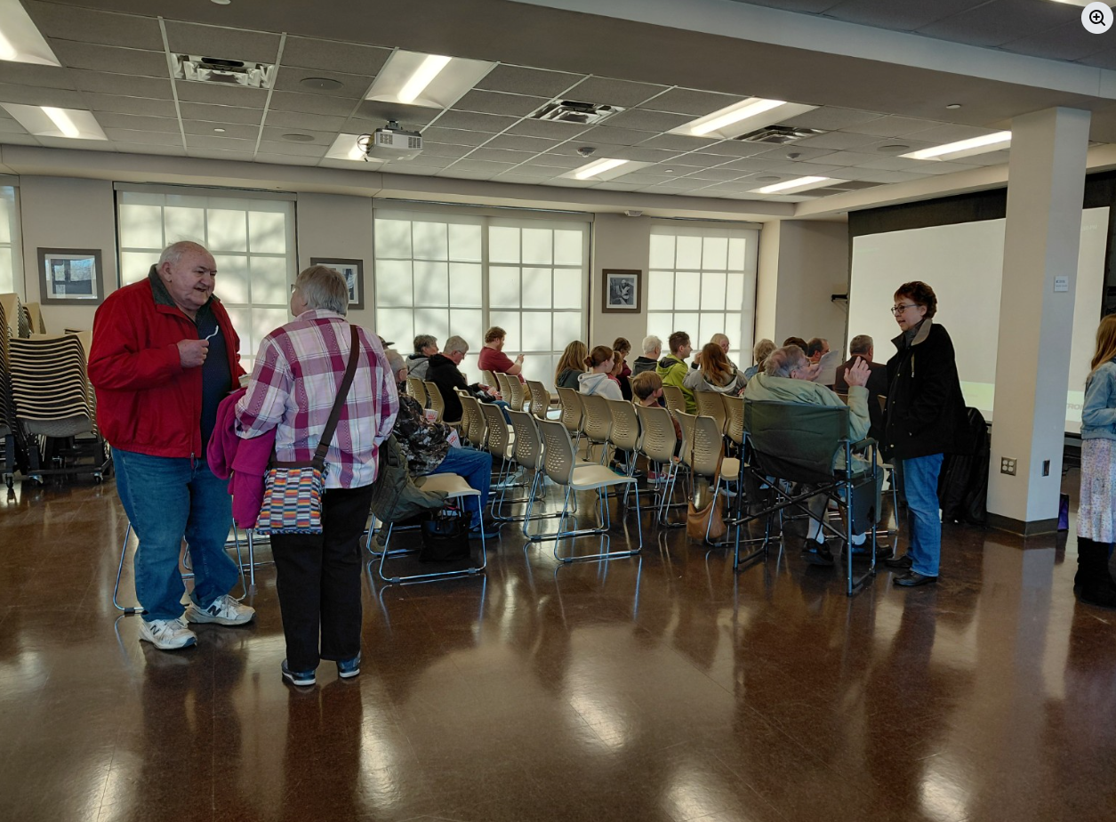People waiting in a community room, some standing and talking, others seated and listening to a presentation on a screen.