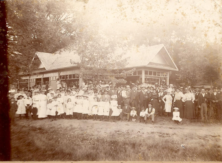 A large group of people, including children and adults, posing outdoors in front of the Riverside Victorian pavilion with trees in the background. The photo appears to be old and sepia-toned.
