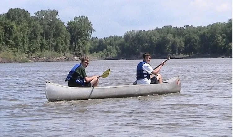 Two young men in a canoe on a calm Minnesota River, paddling and enjoying a sunny day surrounded by trees.