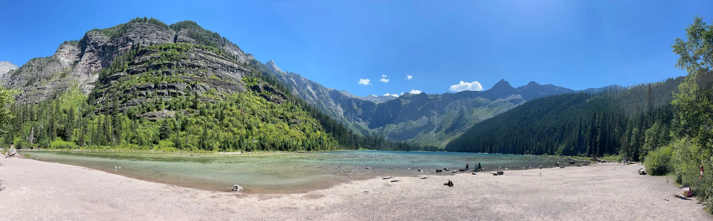 A panoramic view of a serene lake surrounded by lush green trees and towering mountains under a clear blue sky.