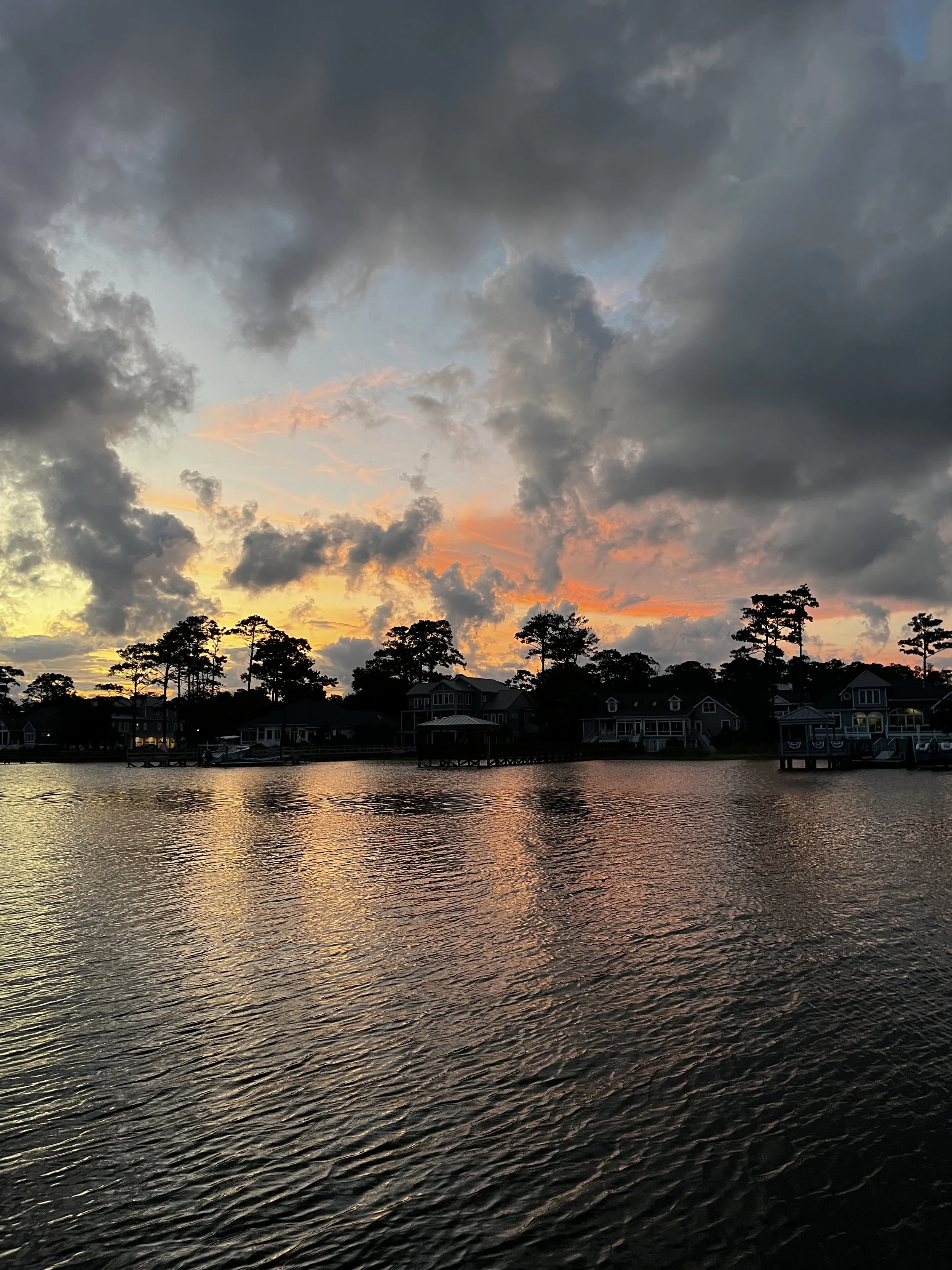 Sunset over a body of water with dark clouds in the sky and houses along the shoreline.