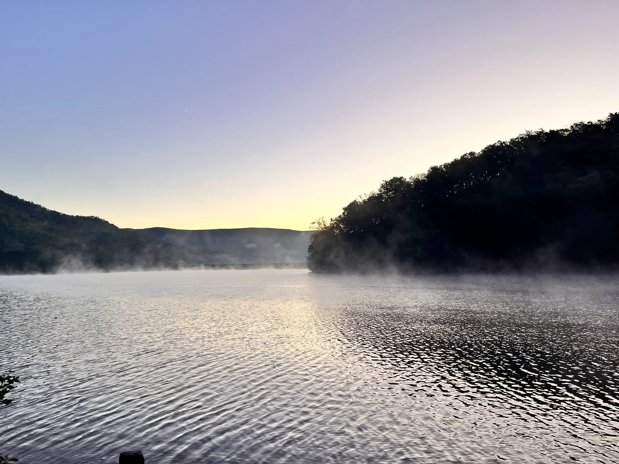 A tranquil lake with mist rising above the water, surrounded by hills and dense trees, under a clear sky at sunrise or sunset.