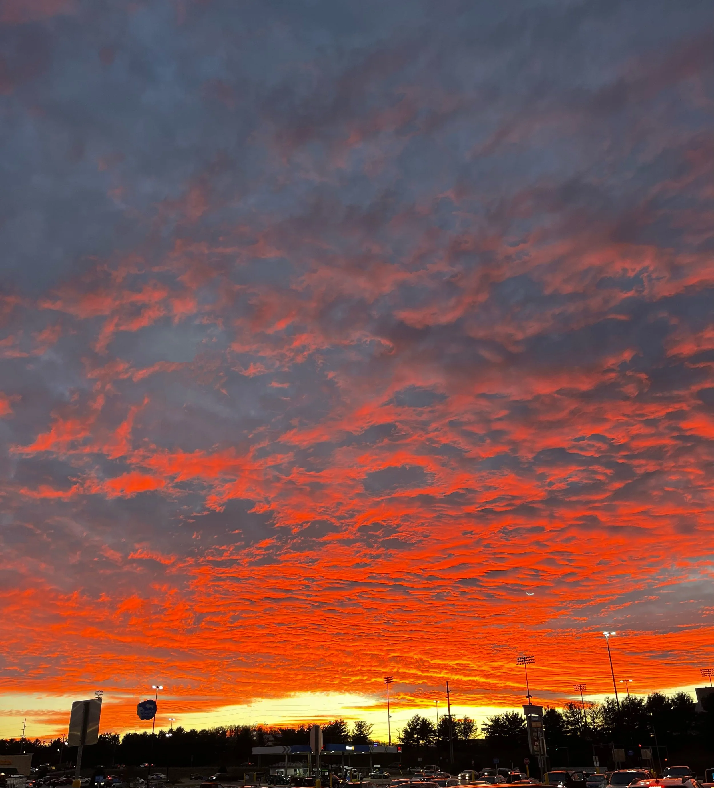 Vibrant sunset sky with orange, pink, and purple clouds over a parking lot with cars and stadium lights in the distance.