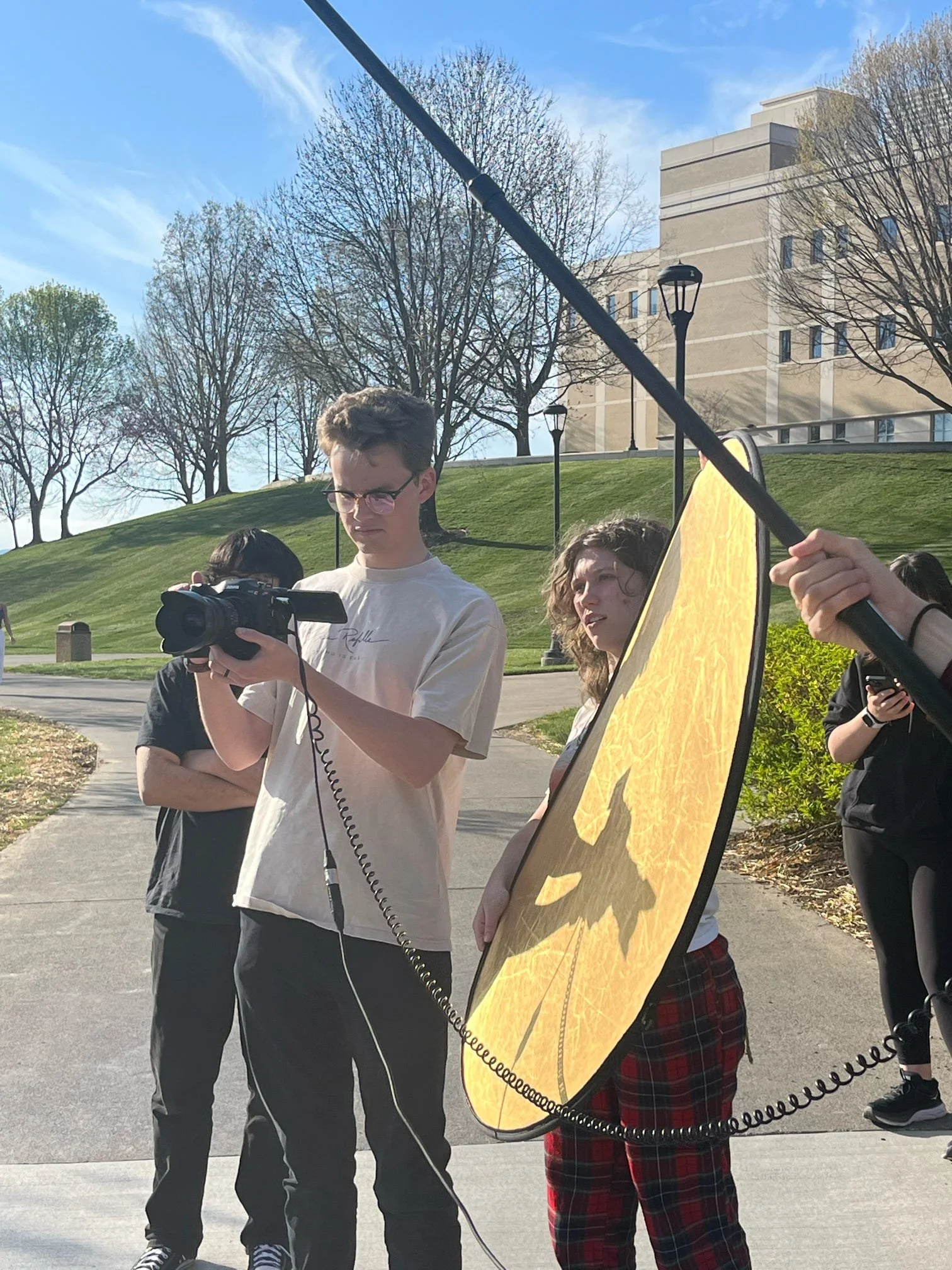 Young adults participating in a video shoot outdoors with a reflector and camera, under a clear blue sky with some clouds and leafless trees.