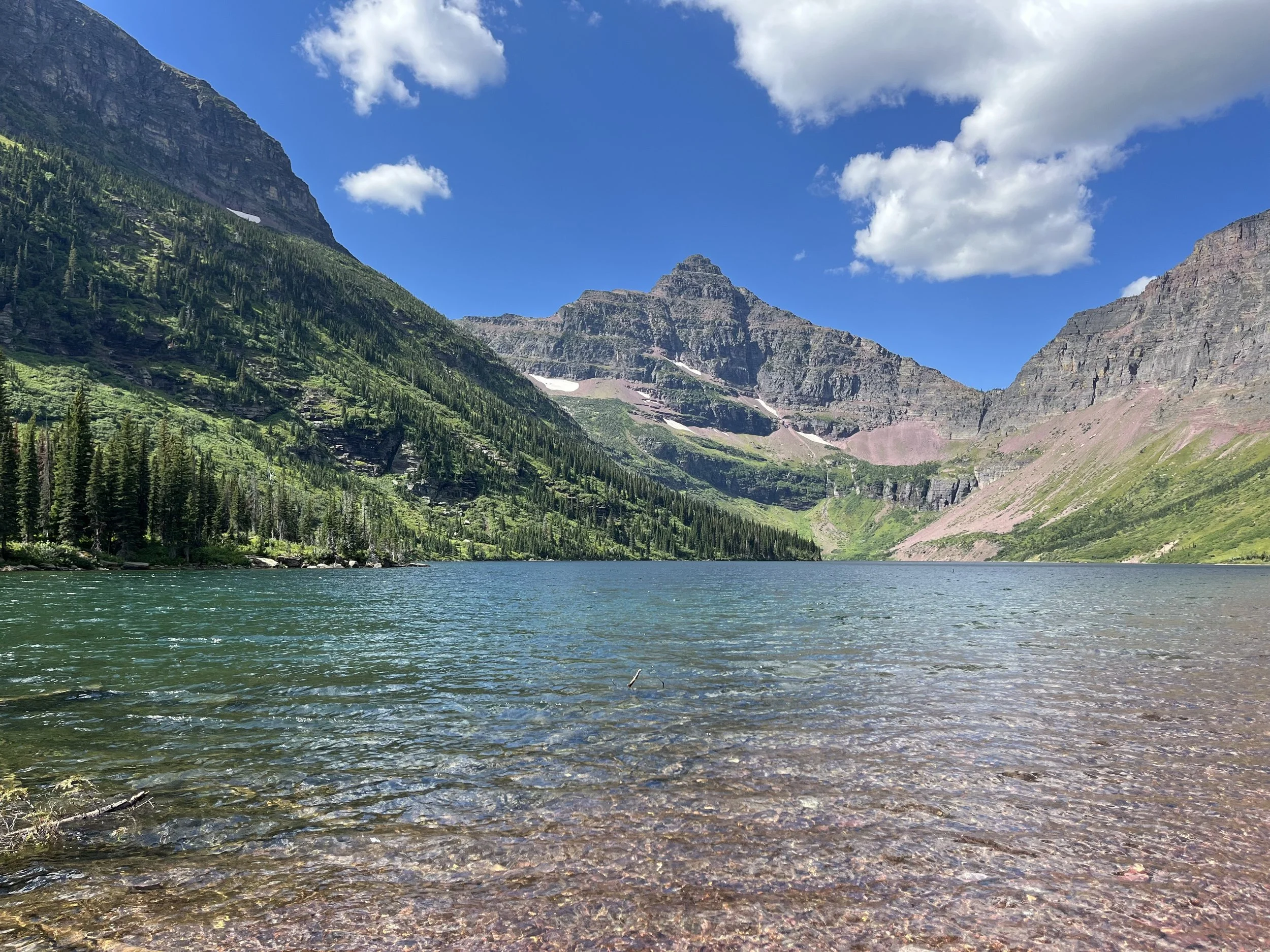 A scenic mountain lake with clear water reflecting the blue sky, surrounded by green pine trees and rugged mountain peaks.