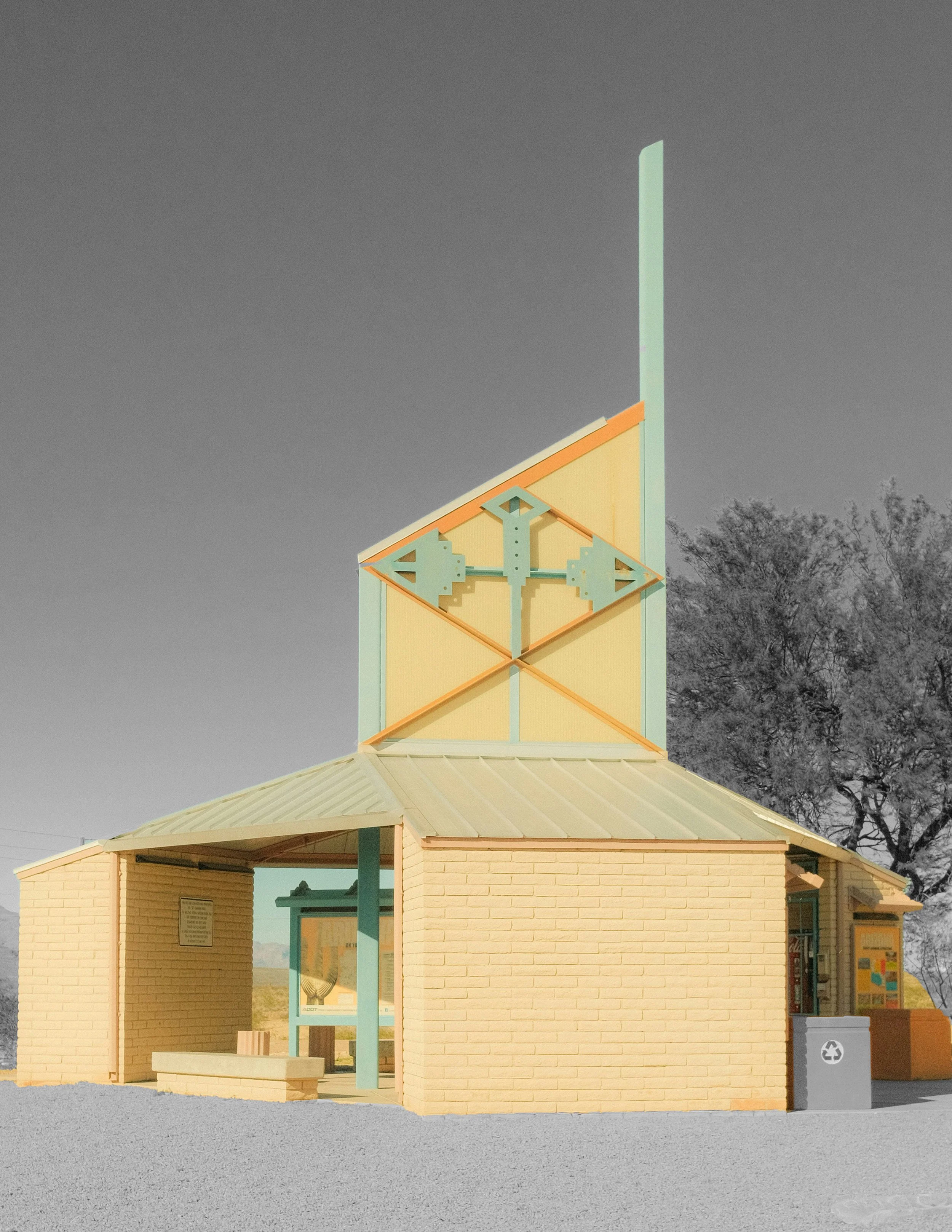 A yellow brick building with a clock tower featuring a pastel green and yellow design, a small covered porch, and a recycling bin nearby, with leafless trees in the background and a clear sky.