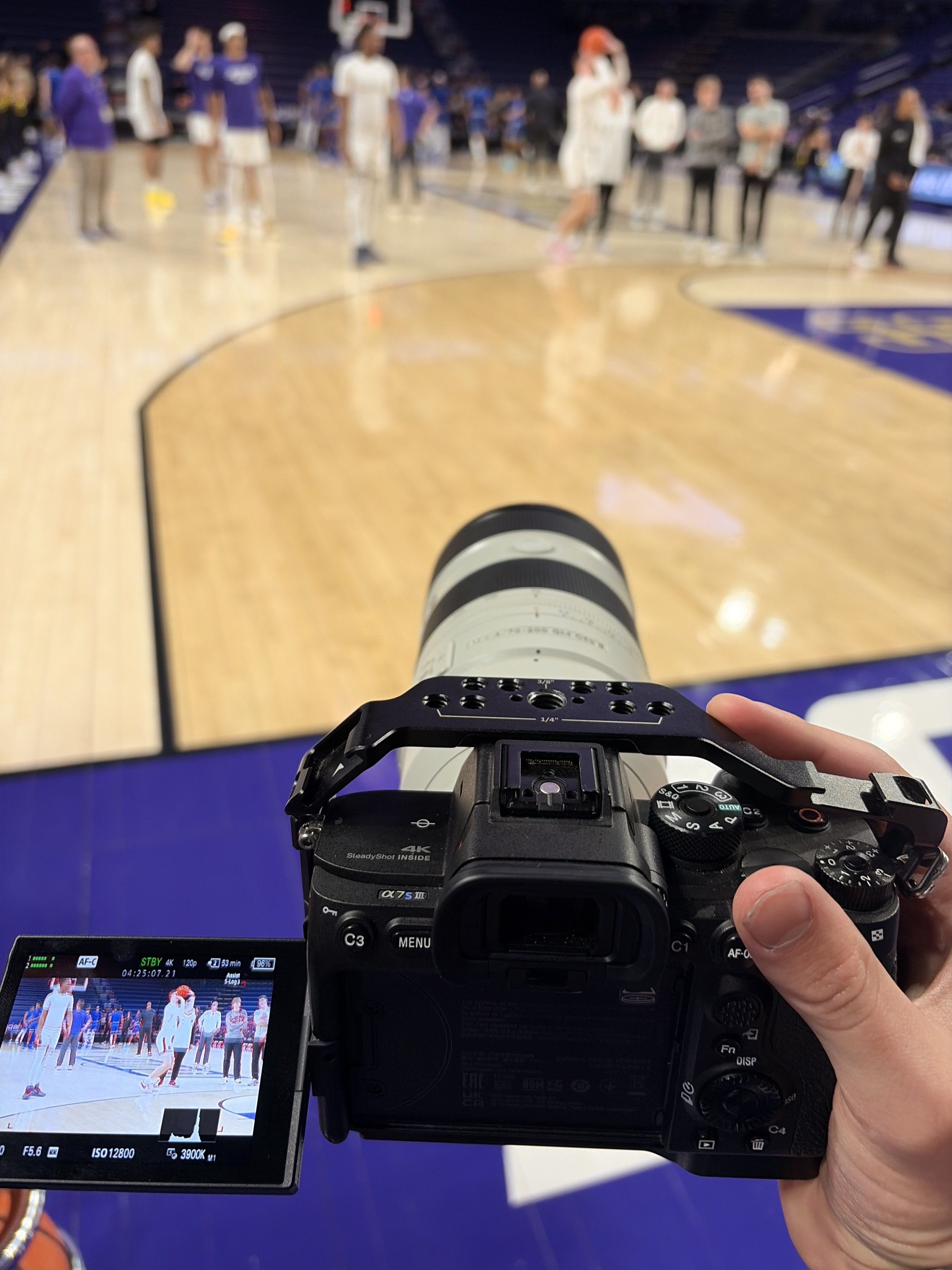 Camera view of a basketball court showing players and officials, with the photographer holding a camera in the foreground.