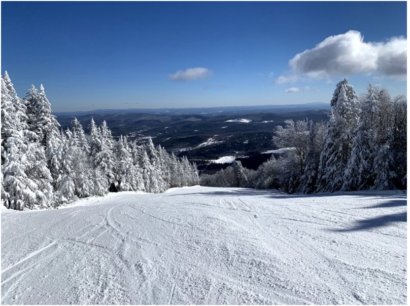 Snow-covered ski slope with frosted trees on both sides and a scenic mountain view in the distance under a partly cloudy blue sky.