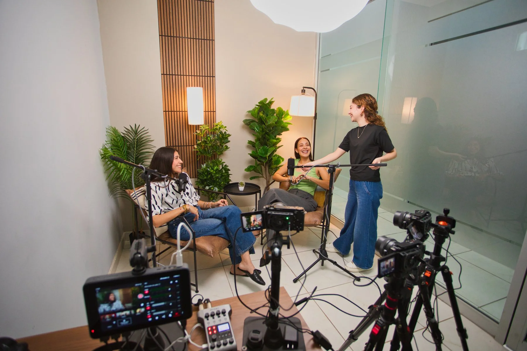 Tres mujeres sentadas en una sala decorada con plantas verdes, mientras una de ellas habla y las otras dos le escuchan y ríen, en un entorno para grabación o entrevista con cámaras y equipo audiovisual.