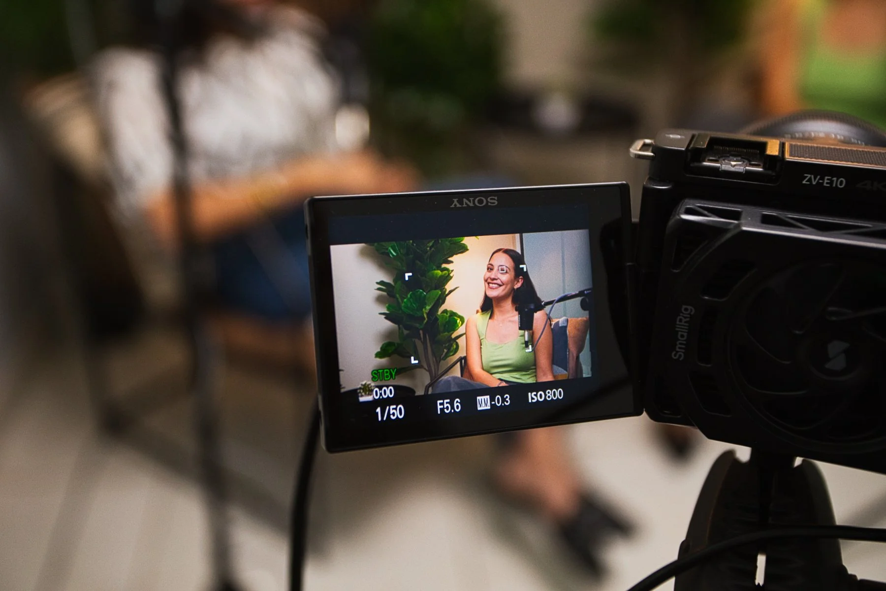 Cuadro de una cámara grabando a una mujer sonriendo, sentada junto a una planta