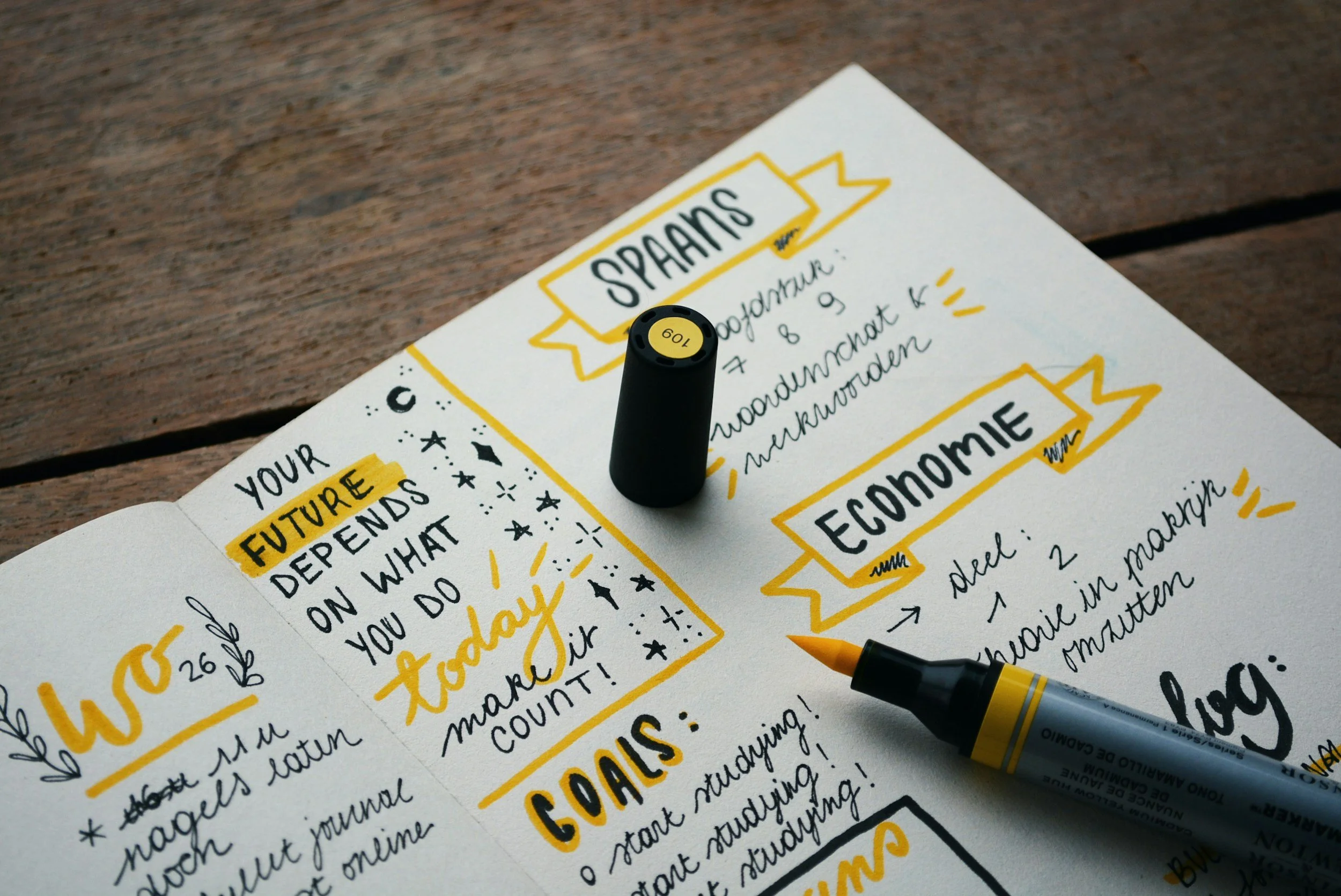 Close-up of a handwritten journal page with yellow and black ink, featuring words and banners such as "SPARKS," "ECONOMIE," and "FUTURES," and a yellow marker pen placed on the page.