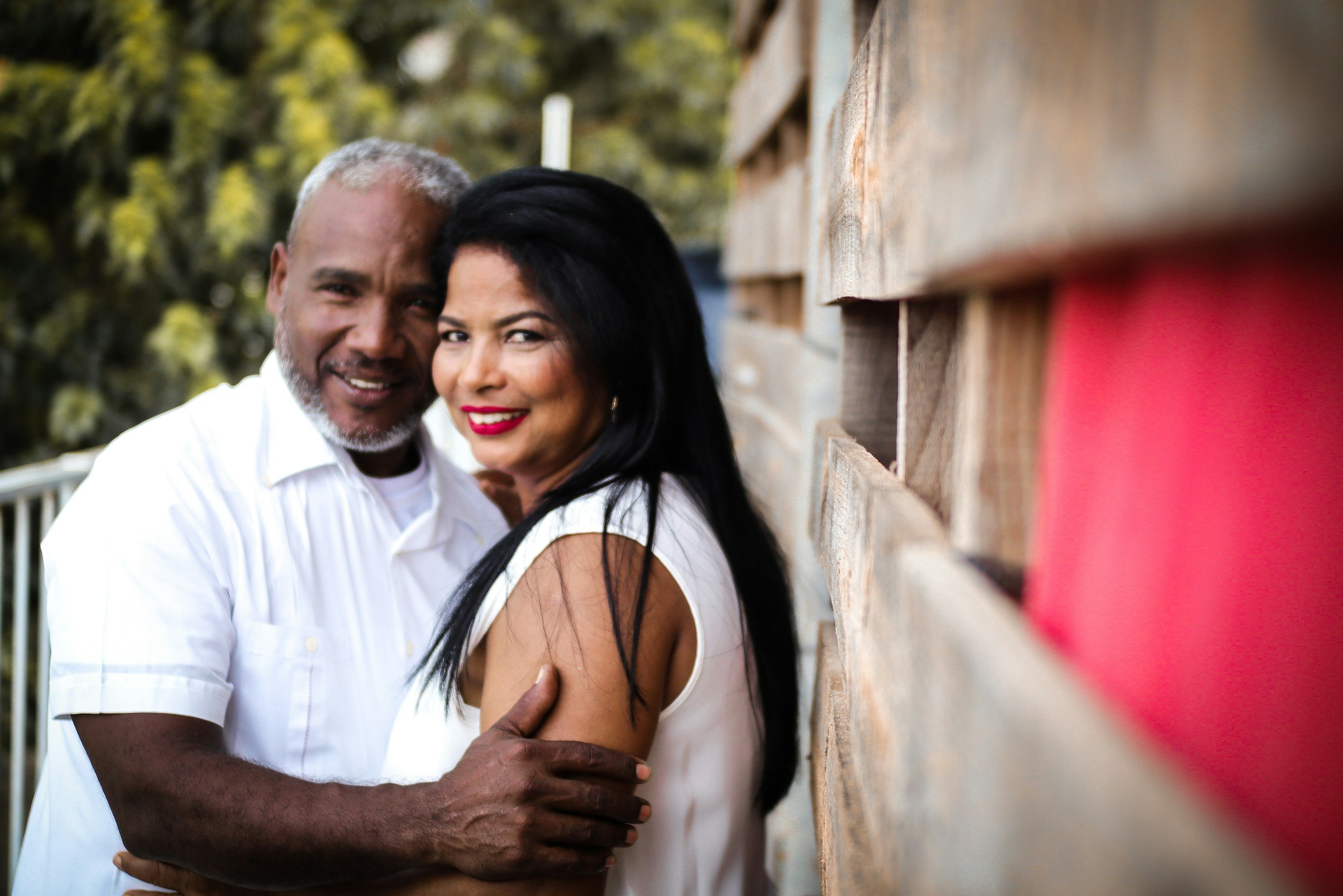 An older man with gray hair and a beard smiling and hugging a woman with long dark hair, standing next to a wooden wall with mailboxes.