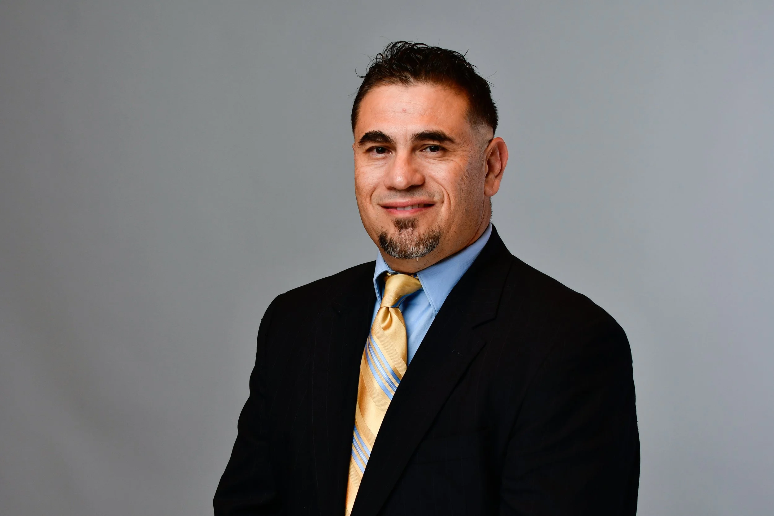 Headshot of a man in a black suit, blue shirt, and yellow striped tie, smiling lightly against a plain gray background.