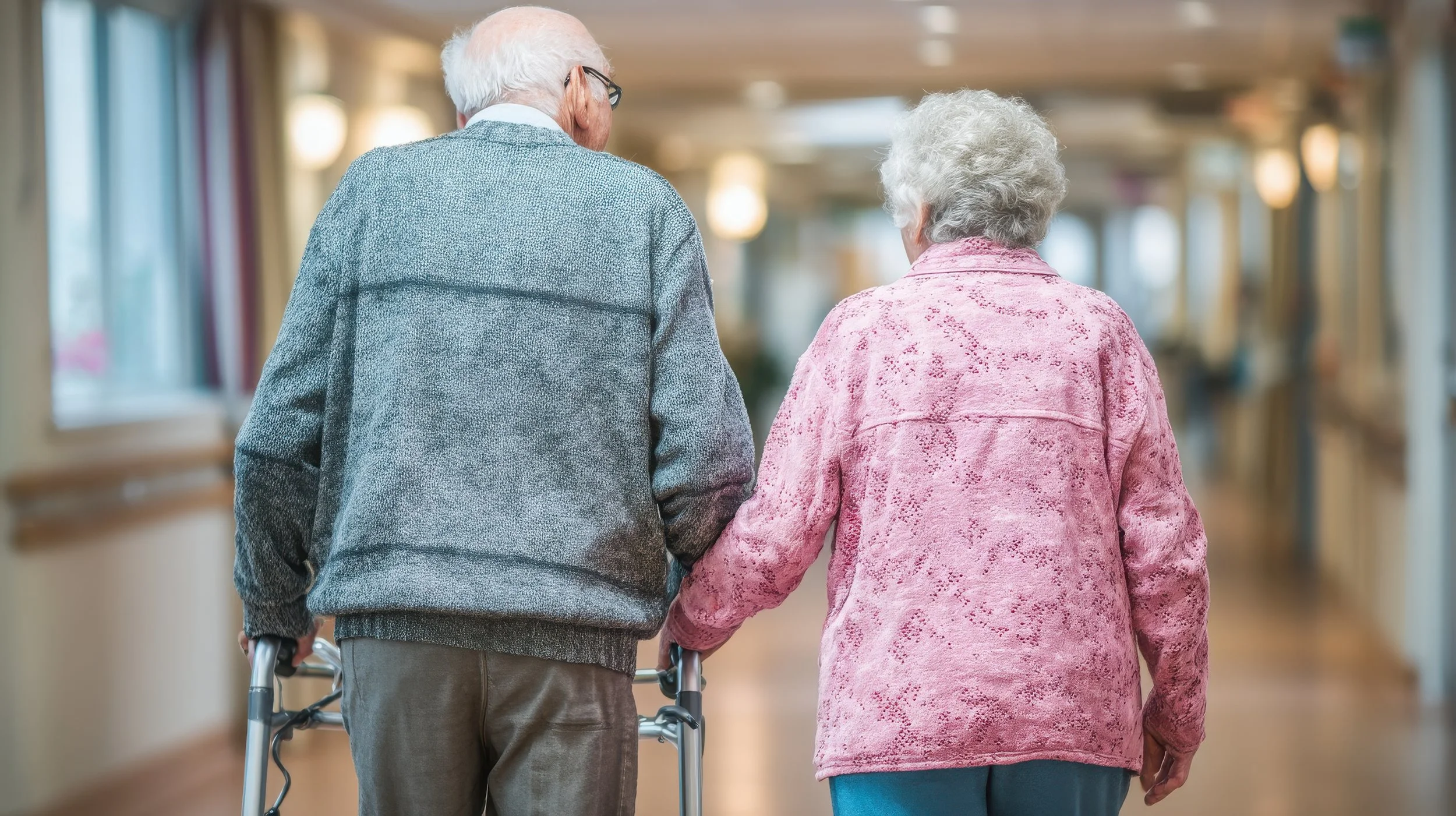 Two elderly women, one with white hair and one with gray hair, walk down a hallway holding hands. The woman on the left uses a walker. The hallway has large windows and warm lighting.