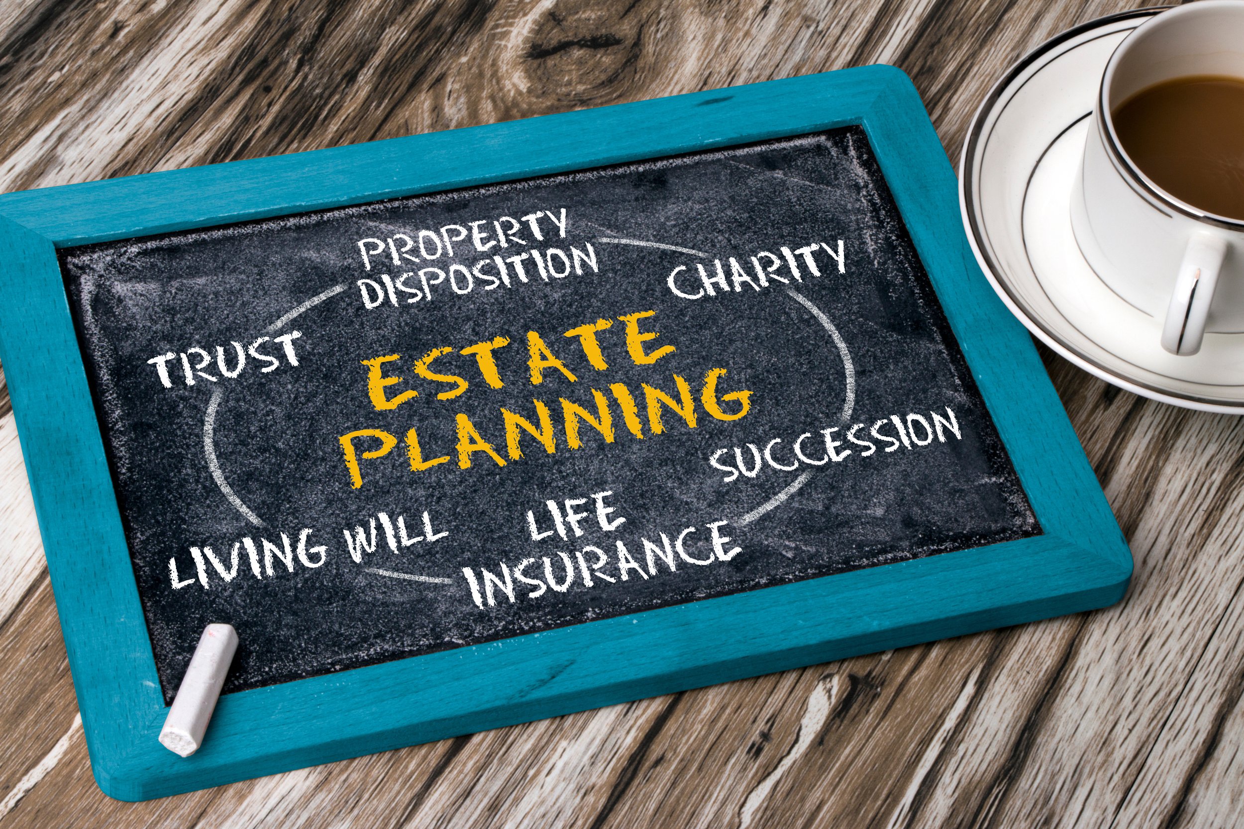 A small chalkboard with a blue frame displaying handwritten words relating to estate planning, including trust, property disposition, charity, will, life insurance, and successin. There is a piece of chalk at the bottom left and a coffee cup on a saucer to the right of the chalkboard, on a wooden surface.