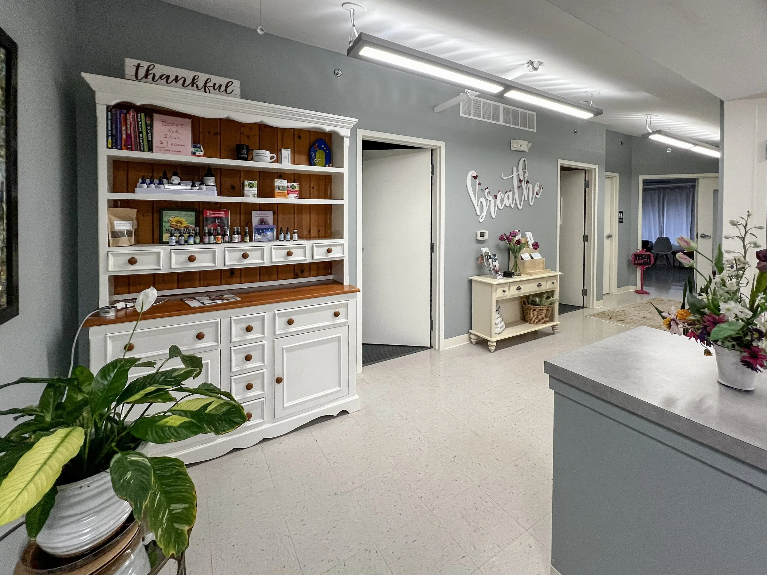 Interior of a building with a white reception desk, a light blue wall, and decorative signs that say 'thankful' and 'be brave'. There are plants, flowers, and a small table with brochures. Overhead lighting and hallway doors are also visible.