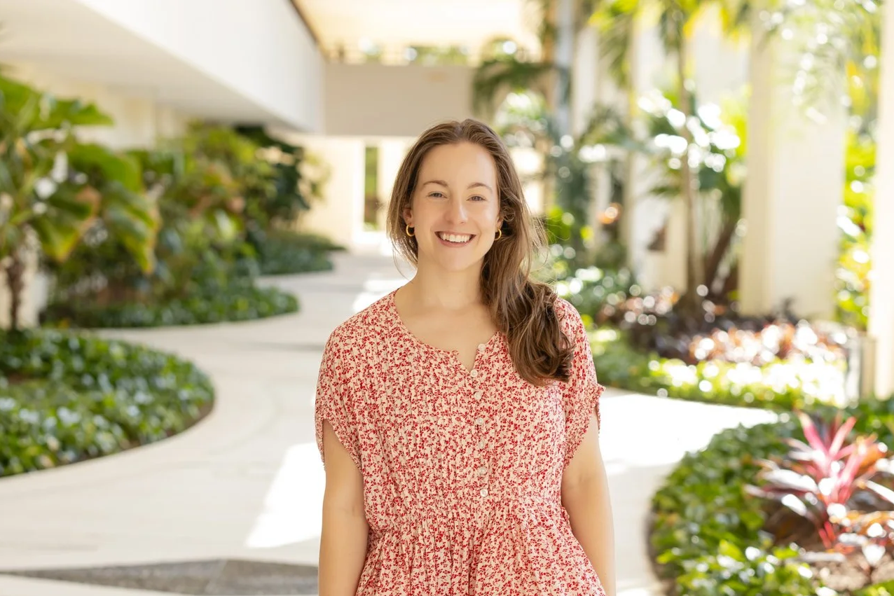 A young woman with long brown hair, wearing a red patterned dress, smiling and standing outdoors in a sunny, lush garden or atrium with greenery and sunlight.