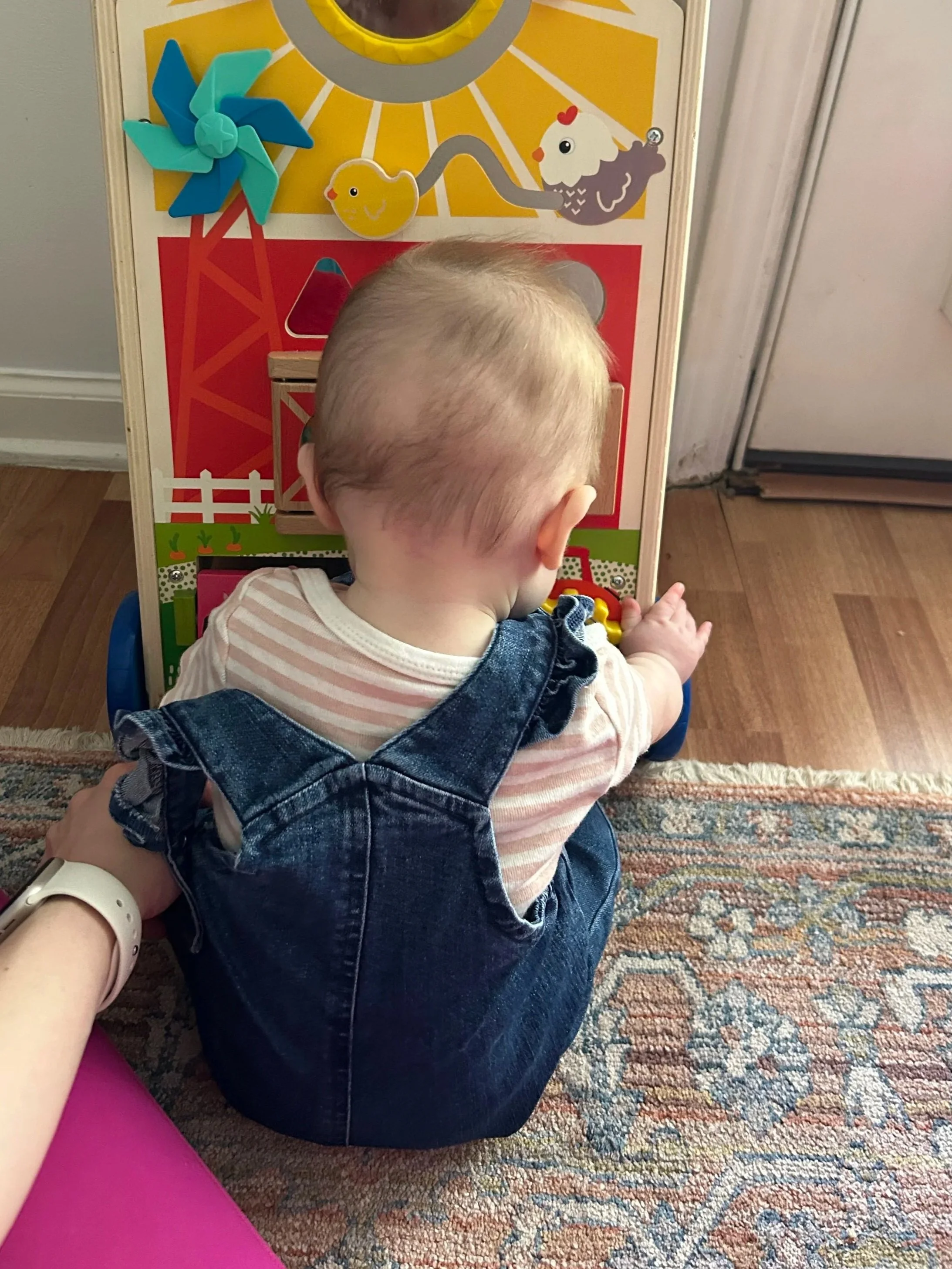 A young child sitting on the floor, playing with a colorful wooden toy barn set. The child's back is to the camera, with short blonde hair and wearing a striped shirt and denim overalls. The floor has a patterned rug, and part of an adult's arm is visible in the lower left corner. The background includes a wall, a door, and a multicolored patterned carpet.