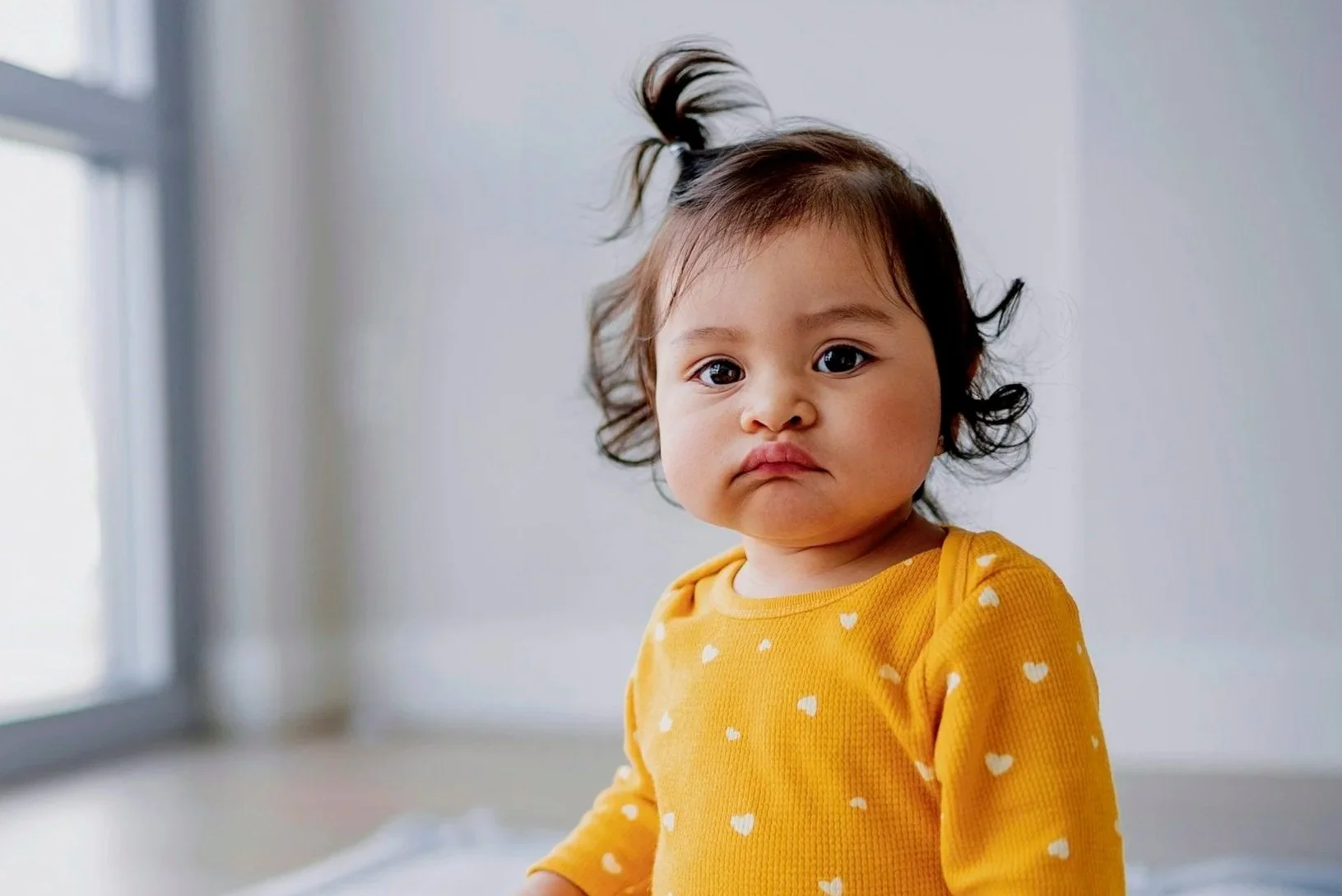 A young girl with curly dark hair in a ponytail, wearing a yellow long-sleeve shirt with white heart patterns, sitting indoors in front of a window with a neutral expression.