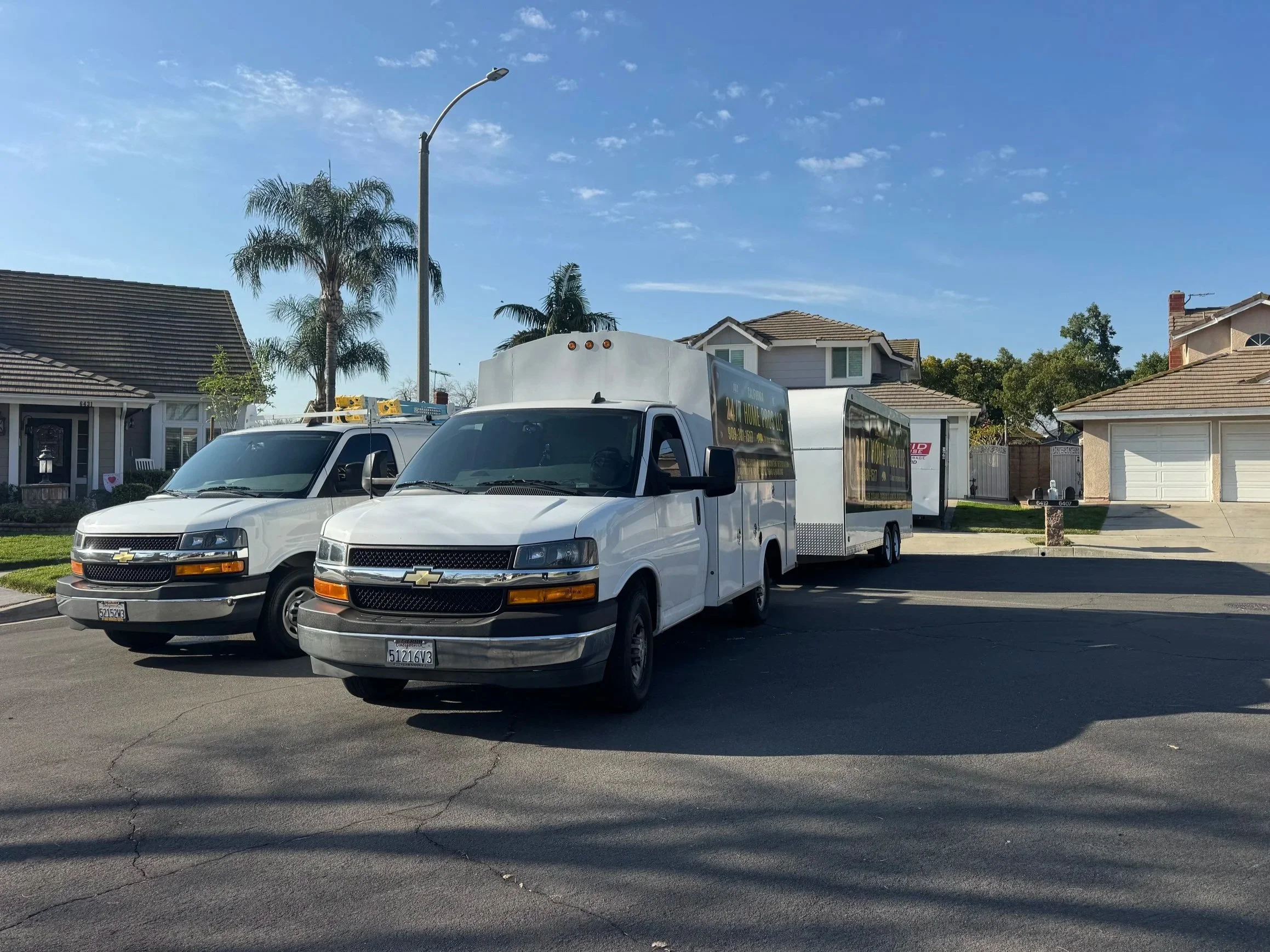 A residential neighborhood street scene with two white service trucks parked in the driveway of a house. The trucks have logo and contact information on the sides. There are houses with tiled roofs, palm trees, and a clear blue sky in the background.