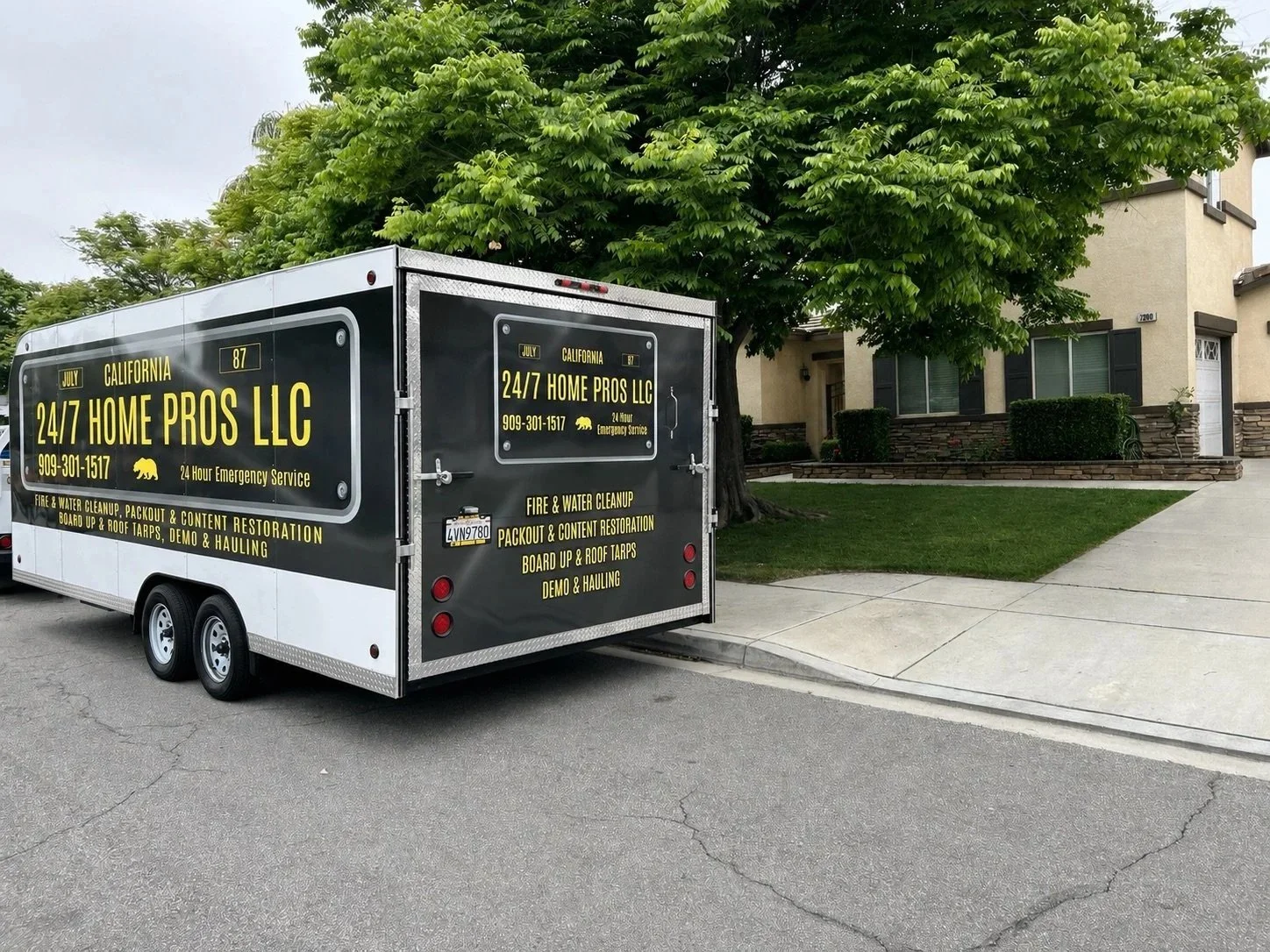 A black and white mobile service trailer parked on a residential street, advertising 24/7 home pros LLC for fire and water cleanup, packout and content restoration, board up and roof tarps, demo and hauling, with a house in the background with a landscaped yard and a tree.