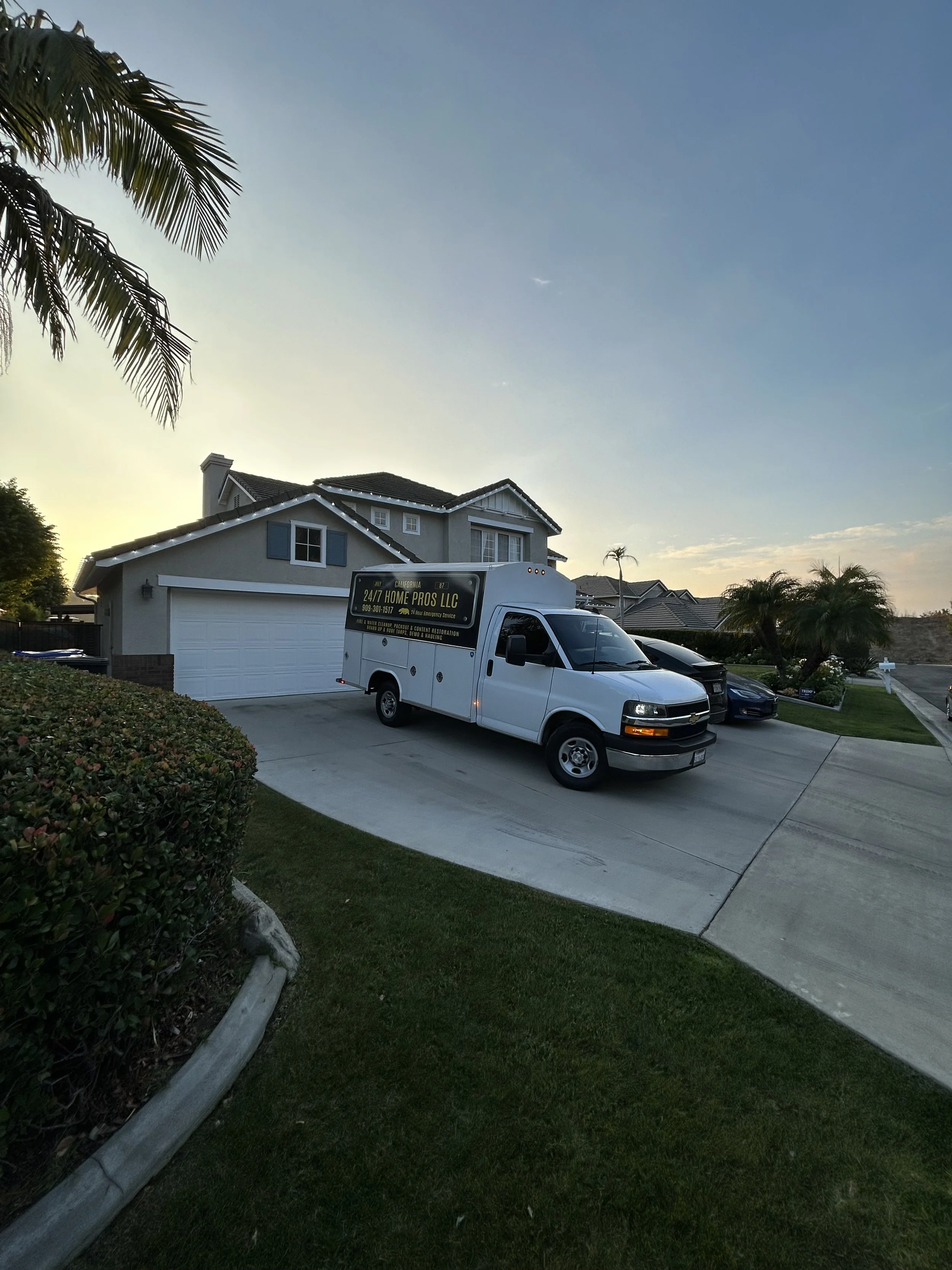 A suburban house with a white garage door and a white work truck parked in the driveway. The truck has a company logo for 24/7 Home Pros LLC. There are palm trees and well-manicured bushes in the yard, and the sky is clear with a slight gradient from yellow to blue at sunset.