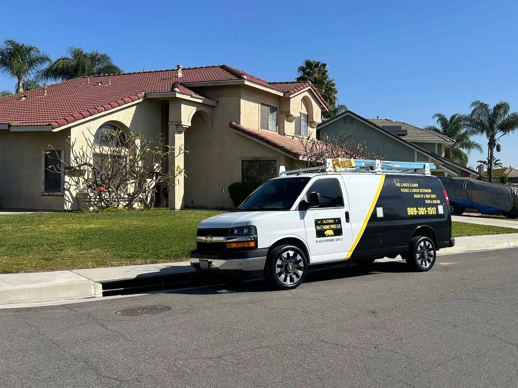 A white and black service van parked in front of a beige house with a red-tiled roof and palm trees in the background, with a clear blue sky overhead.