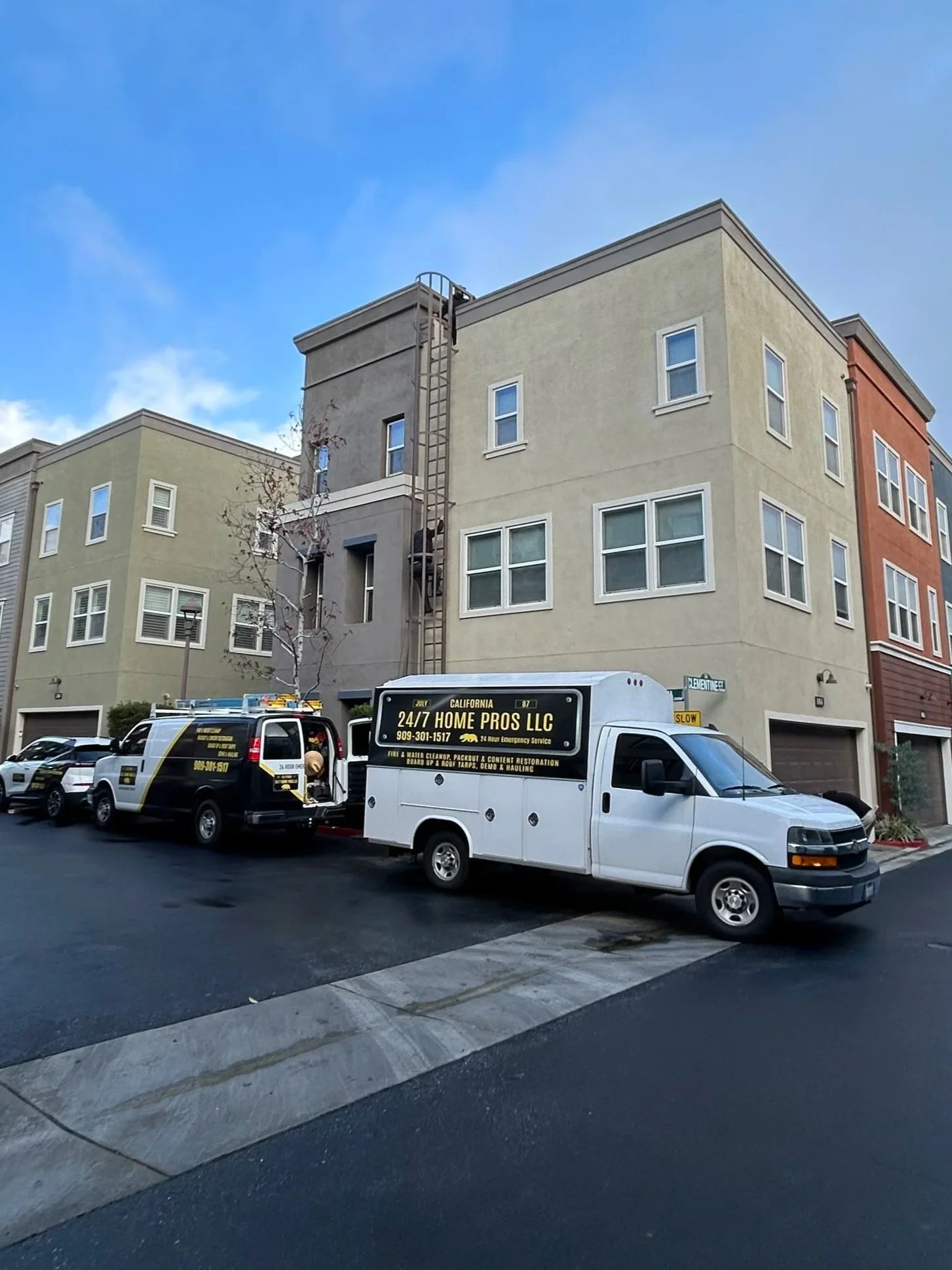 A street view showing parked service vehicles in front of a multi-story residential building with a beige and orange exterior. One vehicle advertises home repair services.