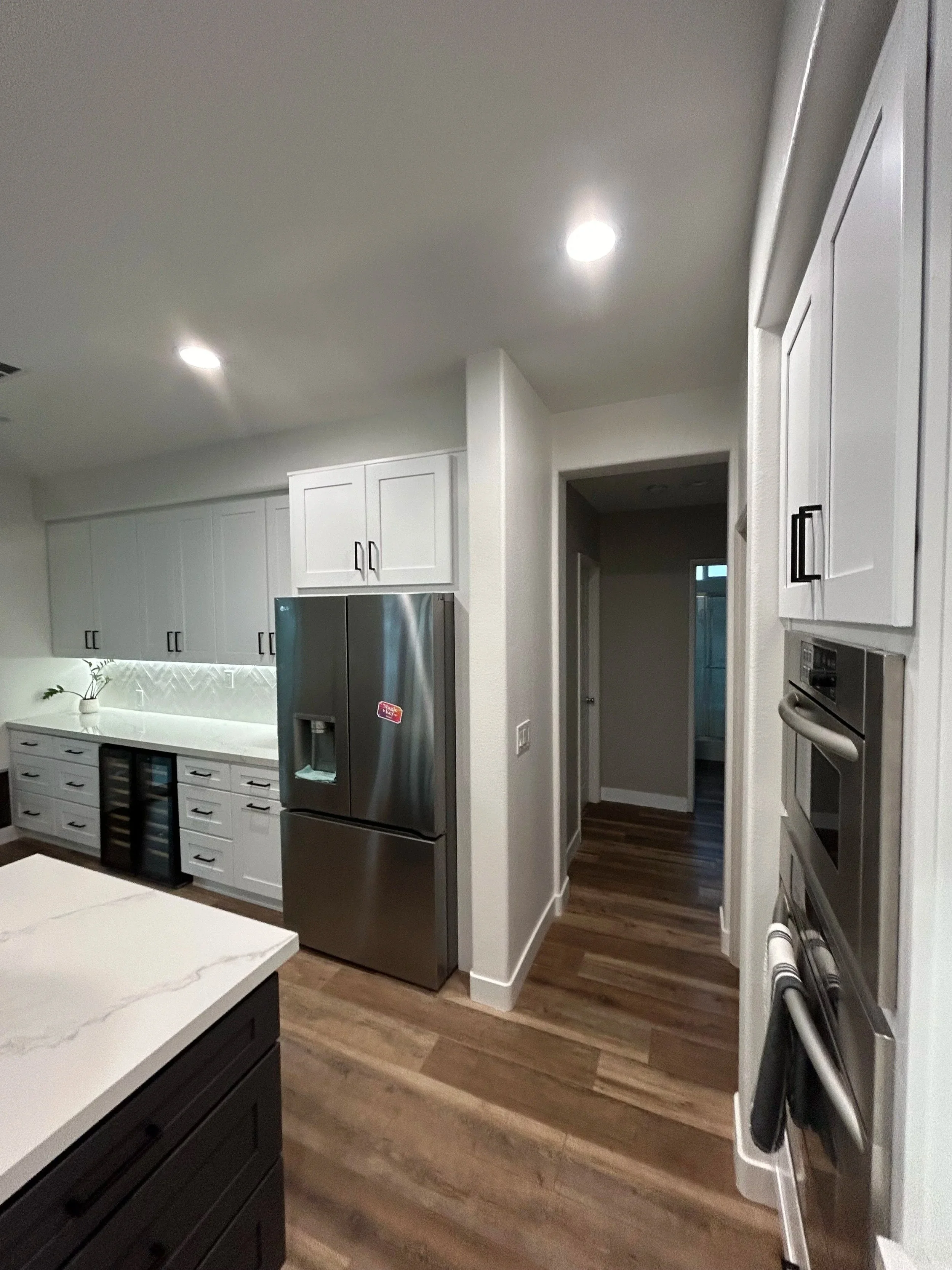 A modern kitchen with white cabinets, stainless steel appliances, a marble-topped island, and wood flooring, illuminated by ceiling lights.