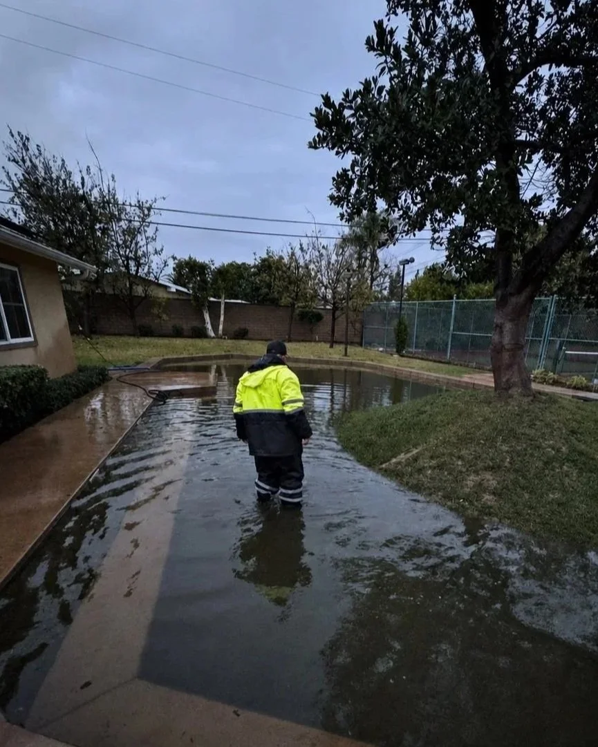 A person in a yellow and black rain jacket standing in ankle-deep water in a backyard with flooded grass, trees, and a patio, during cloudy weather.