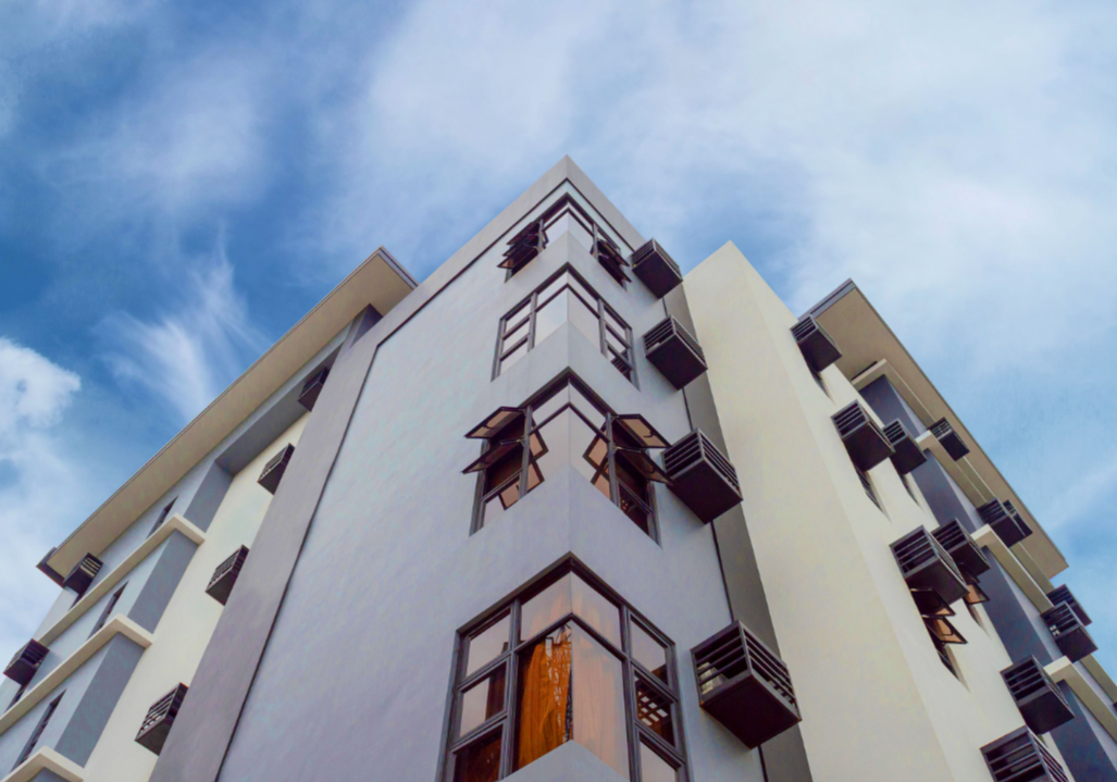 Low-angle view of a modern multi-story apartment building with balconies and large windows, set against a blue sky with some clouds.