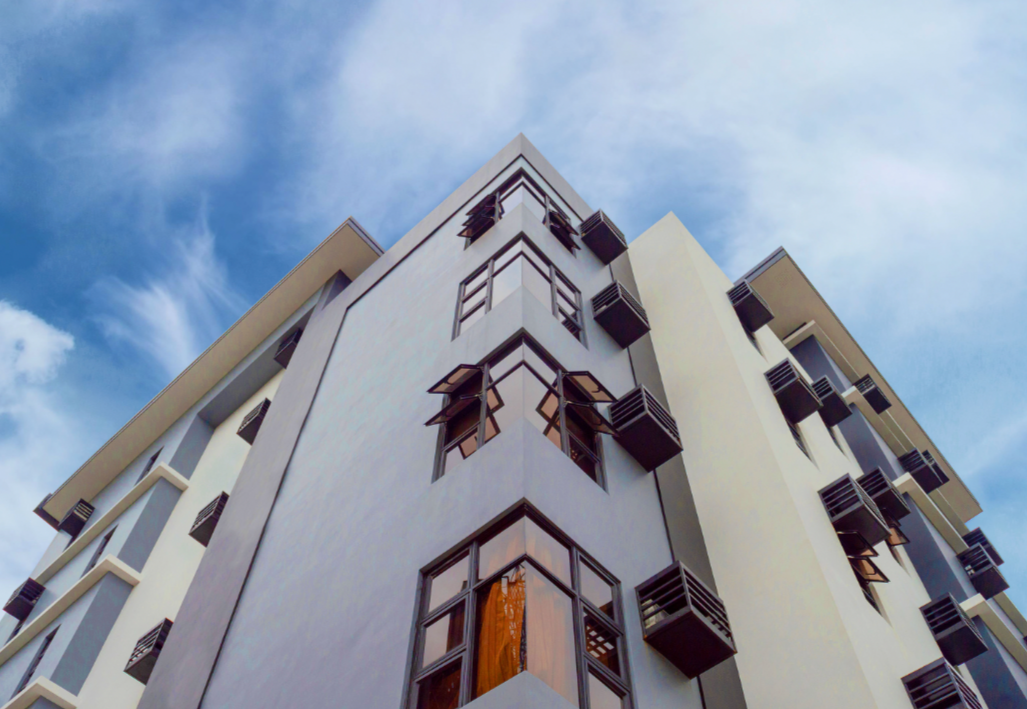 Low-angle view of a modern multi-story apartment building with numerous balconies and large windows, set against a blue sky with scattered clouds.