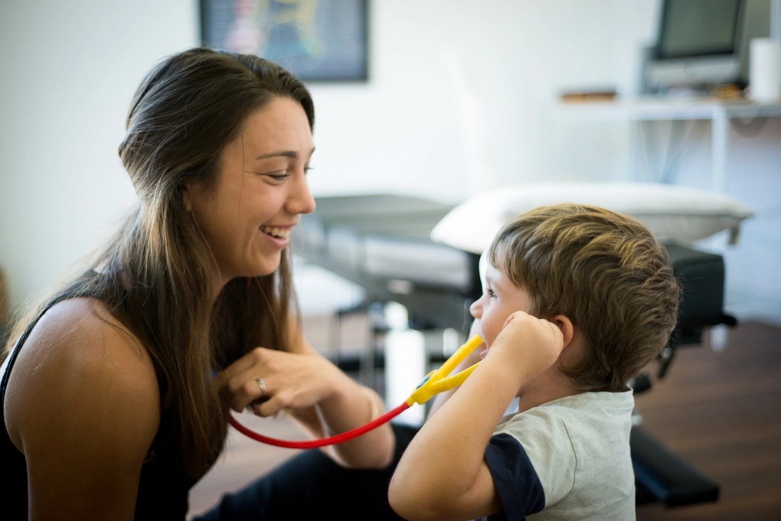 A woman with long brown hair smiling while holding a stethoscope to a young boy's chest in a medical office.