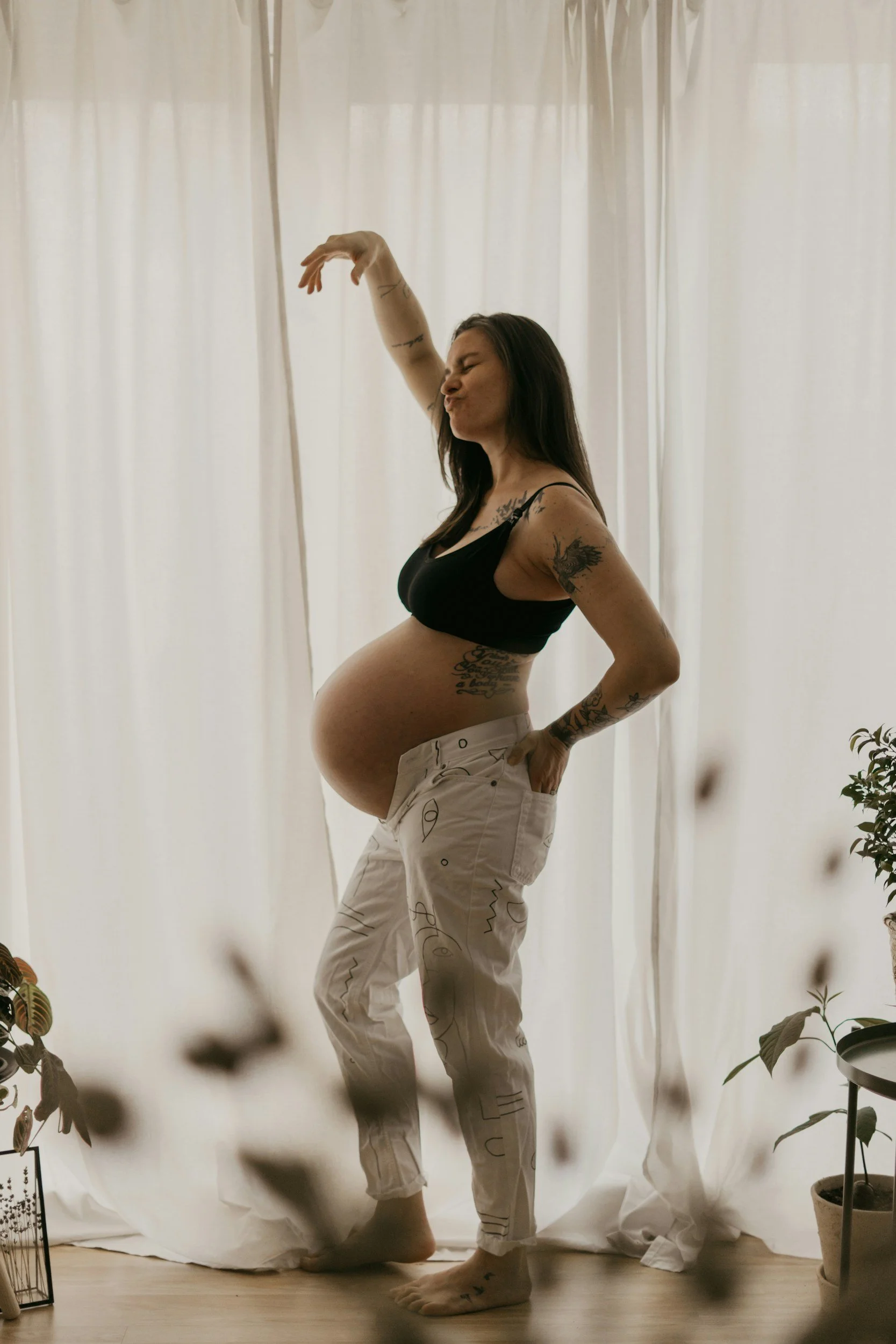 A pregnant woman in a black tank top and white patterned pants standing in front of white curtains, making a playful face with her eyes closed and lips puckered, with plants visible on either side.