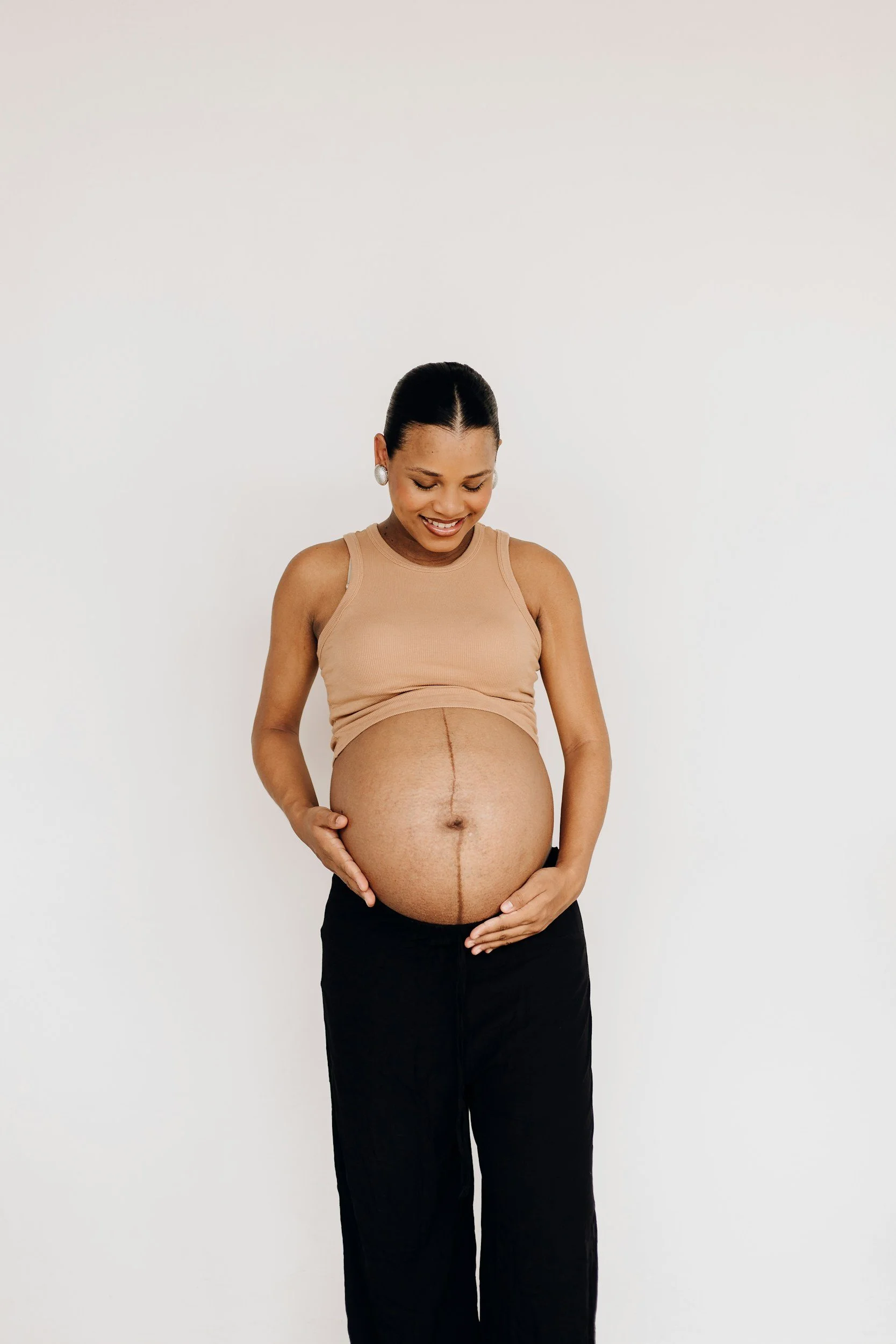 A pregnant woman smiling and looking down, holding her belly with both hands, standing against a plain white background.