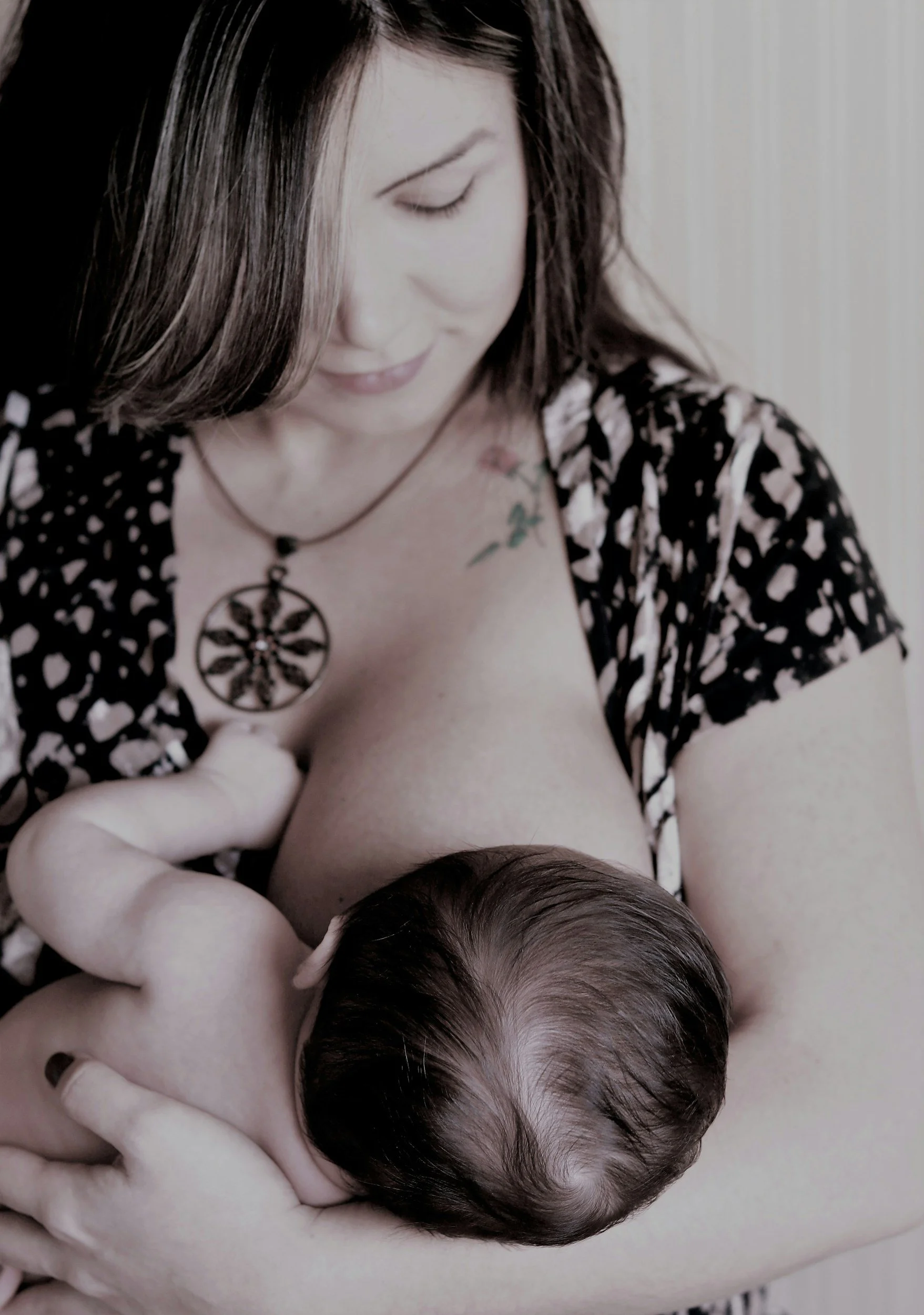 A woman with dark hair and tattoos, wearing a black and white patterned dress and a pendant necklace, is holding a newborn baby with dark hair close to her chest, both looking down.
