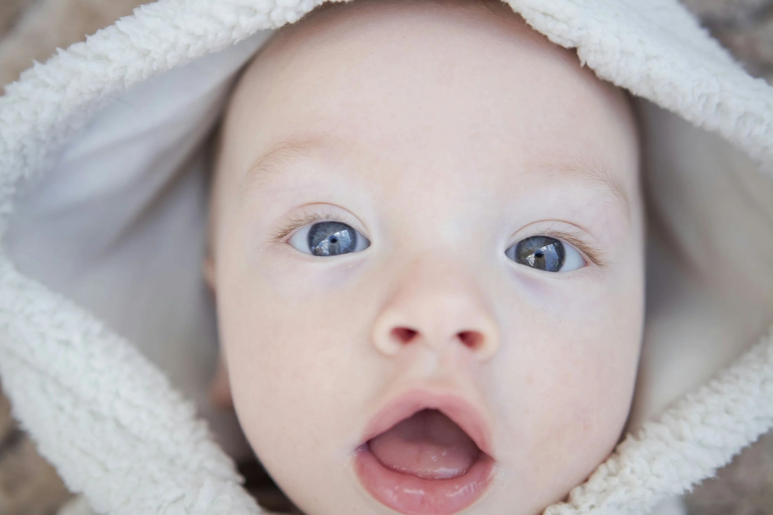 Close-up of a baby's face with blue eyes, open mouth, and fluffy white hood around the head.
