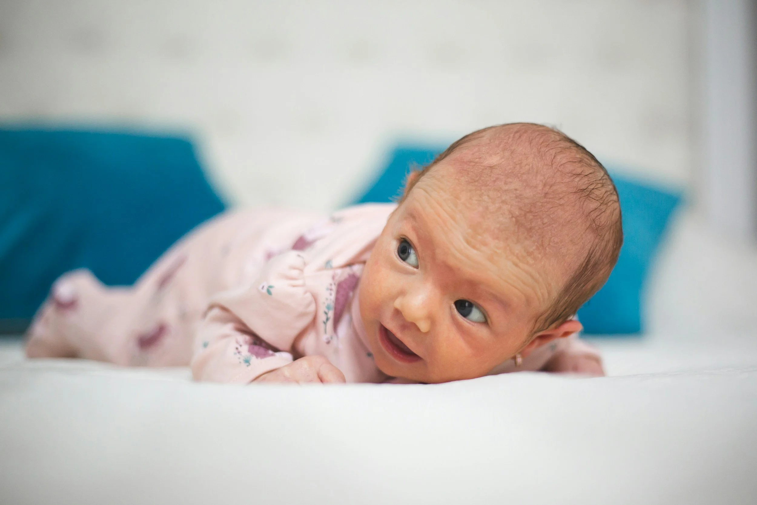 A baby lying on their stomach on a white surface, looking to the right with wide eyes and a slight smile.