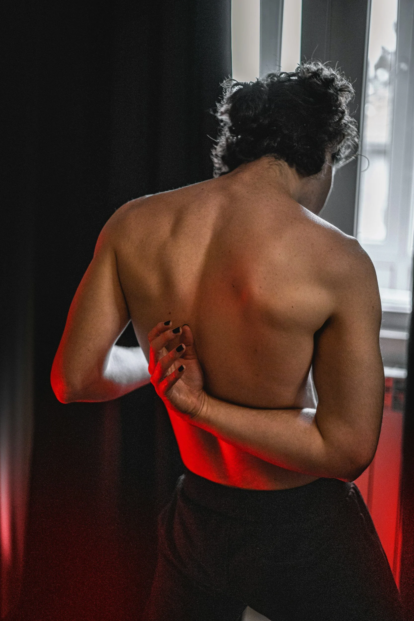 A shirtless person with curly dark hair standing near a window with light coming through, with their back turned. They are holding their left arm with their right hand, which has painted nails. The photo features a dark background with a red glow on the lower left side.